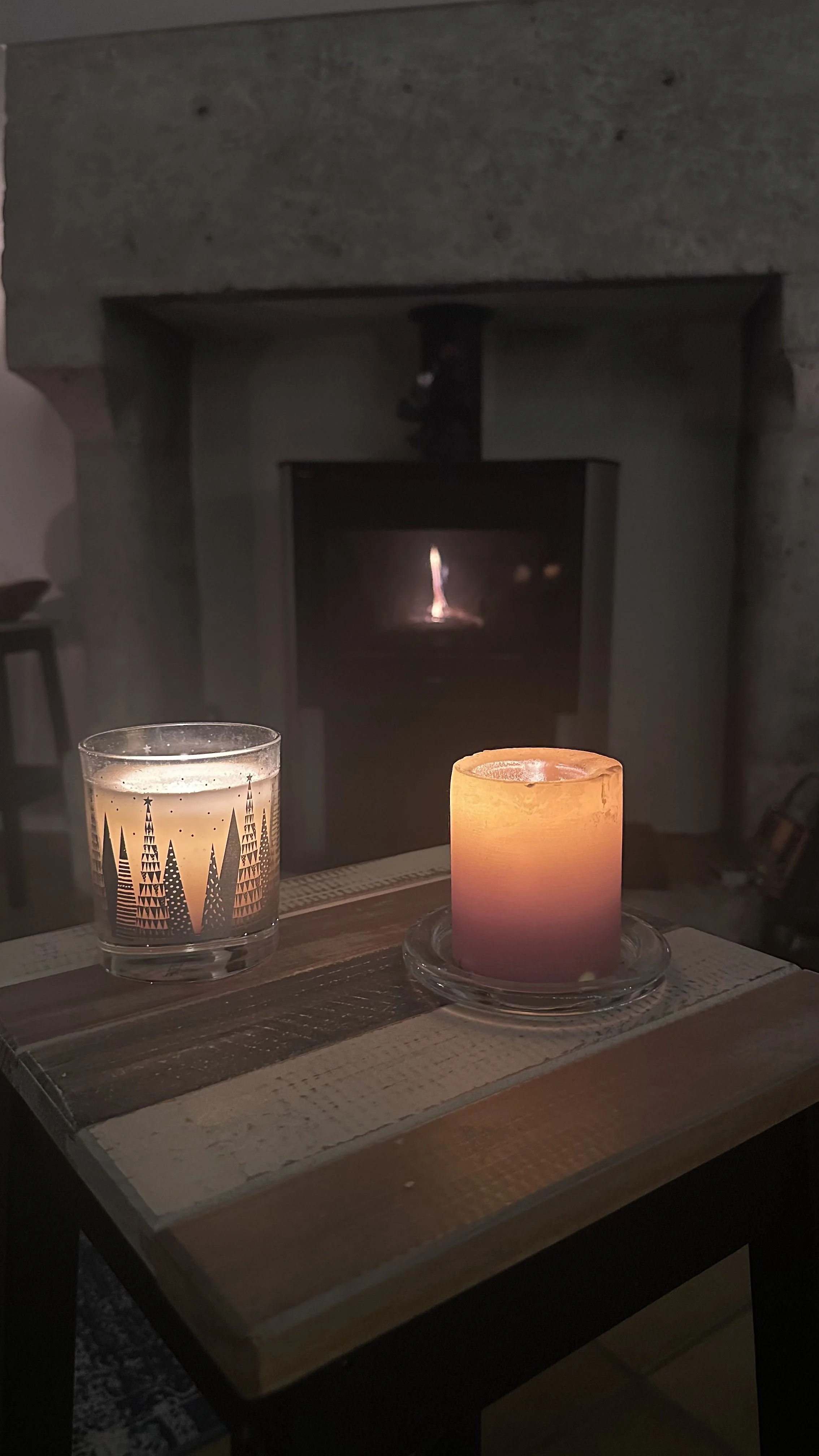 A lit pink candle and a glass of a beverage with black geometric patterns on the outside, placed on a wooden table with a background of a fireplace with a small flame.