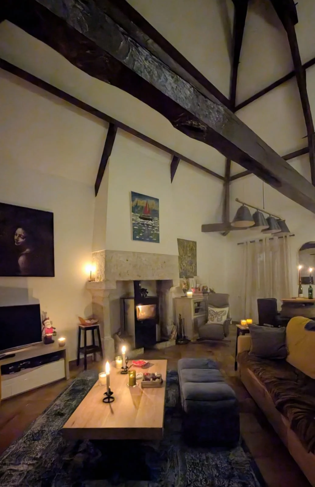 Cozy living room with a lit fireplace, wooden beams on the ceiling, a large sofa, armchair, coffee table with candles, and wall art.