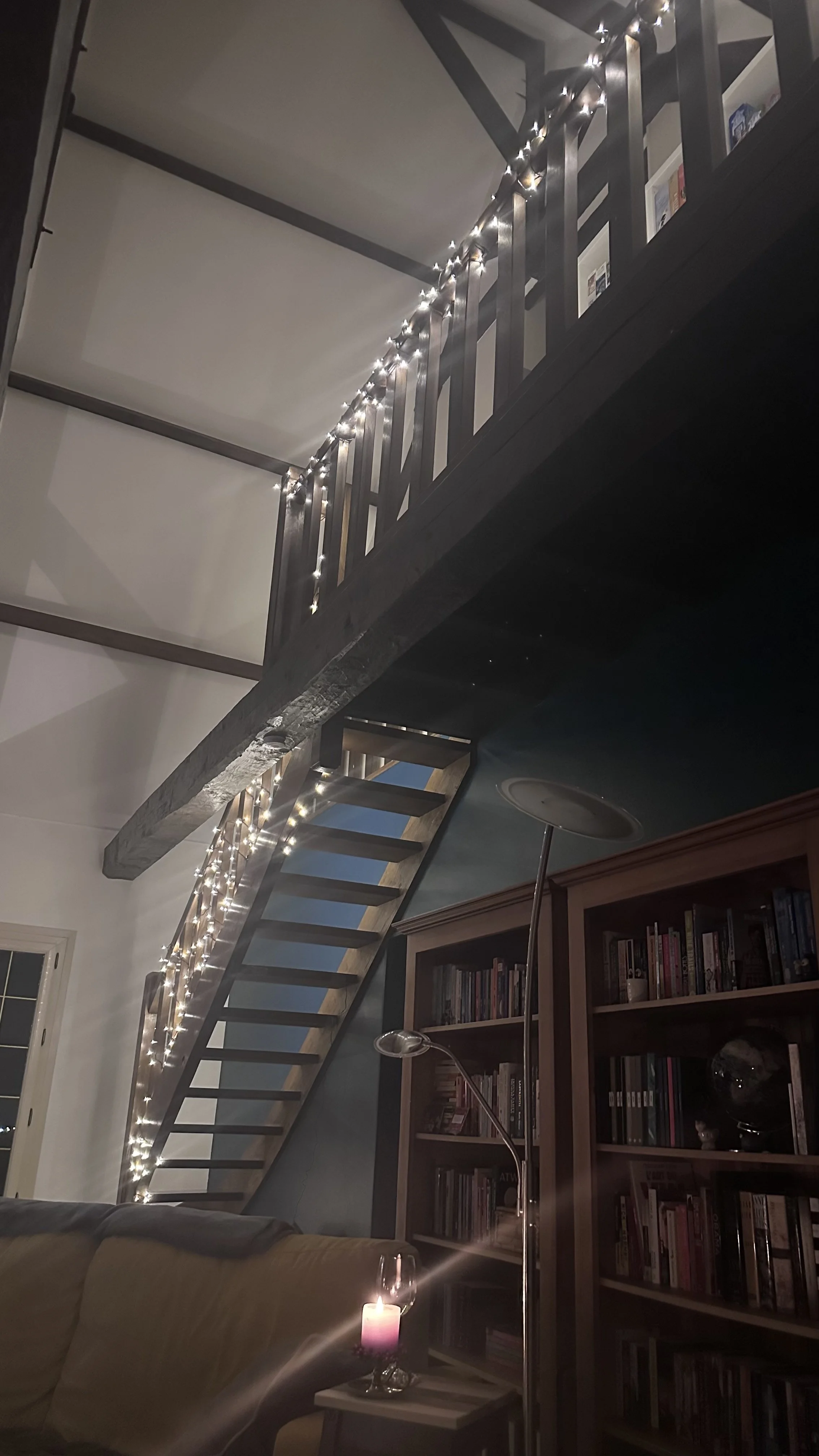 Living room with a couch, a bookshelf filled with books, a small table with a lit candle, and a staircase decorated with string lights leading to a loft area.