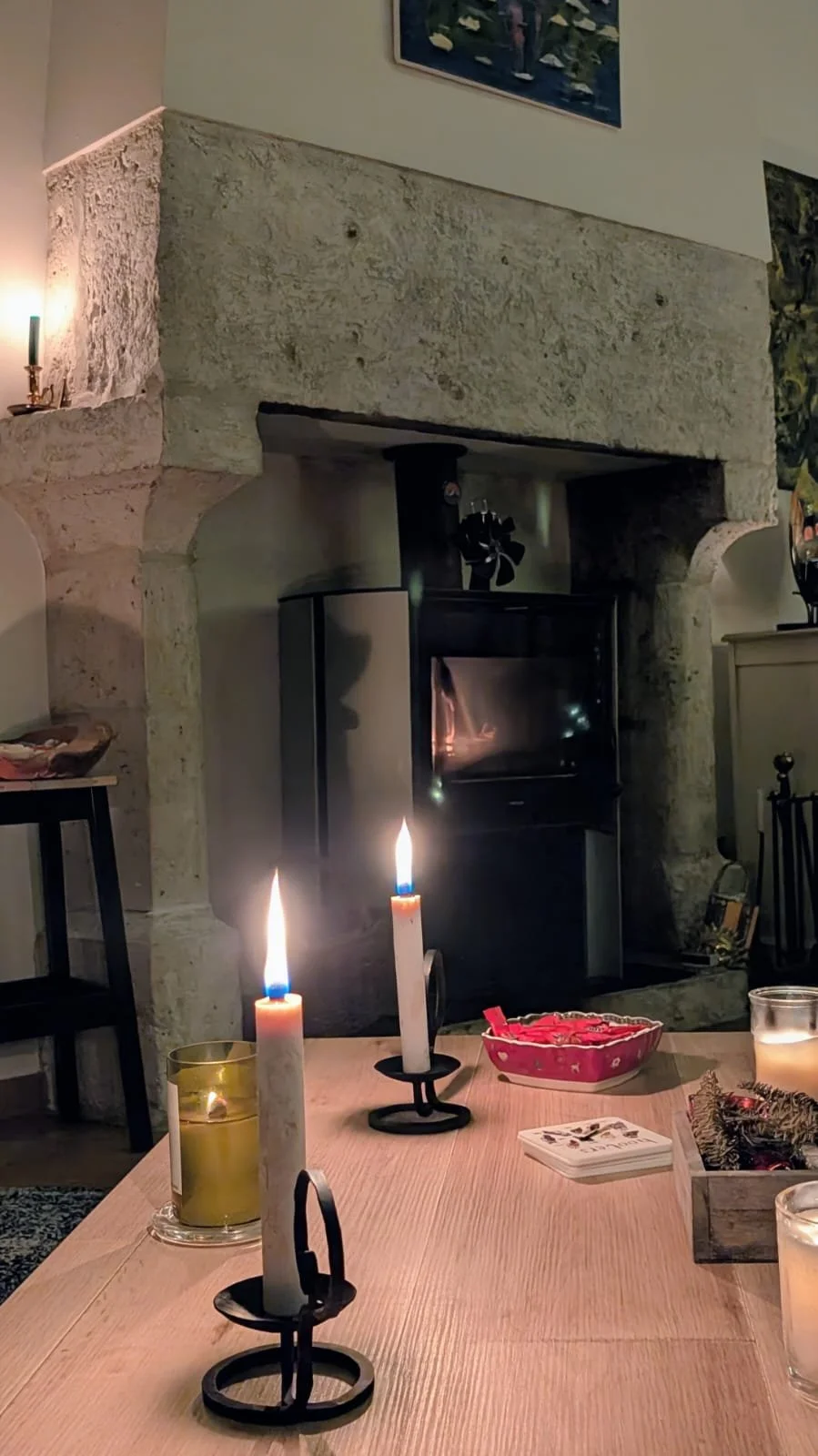 A cozy living room with a stone fireplace, lit candles on a wooden table, and some colorful decorations, creating a warm atmosphere.