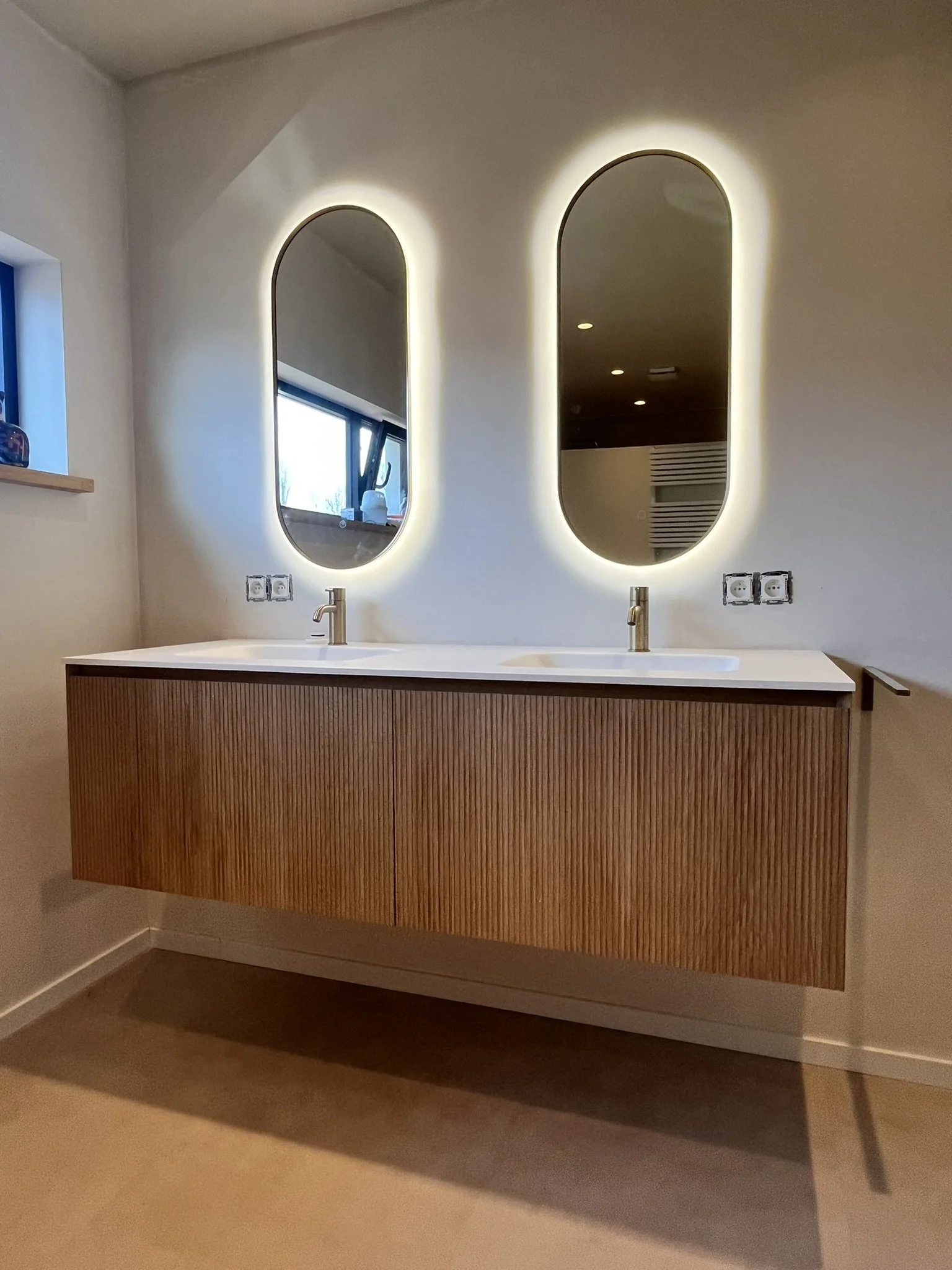 Een moderne badkamer met twee ronde spiegels met achtergrondverlichting, een houten wastafel onder twee wastafelkranen, boven een witte muur, met een kleine raam met een vensterbank aan de linkerzijde.