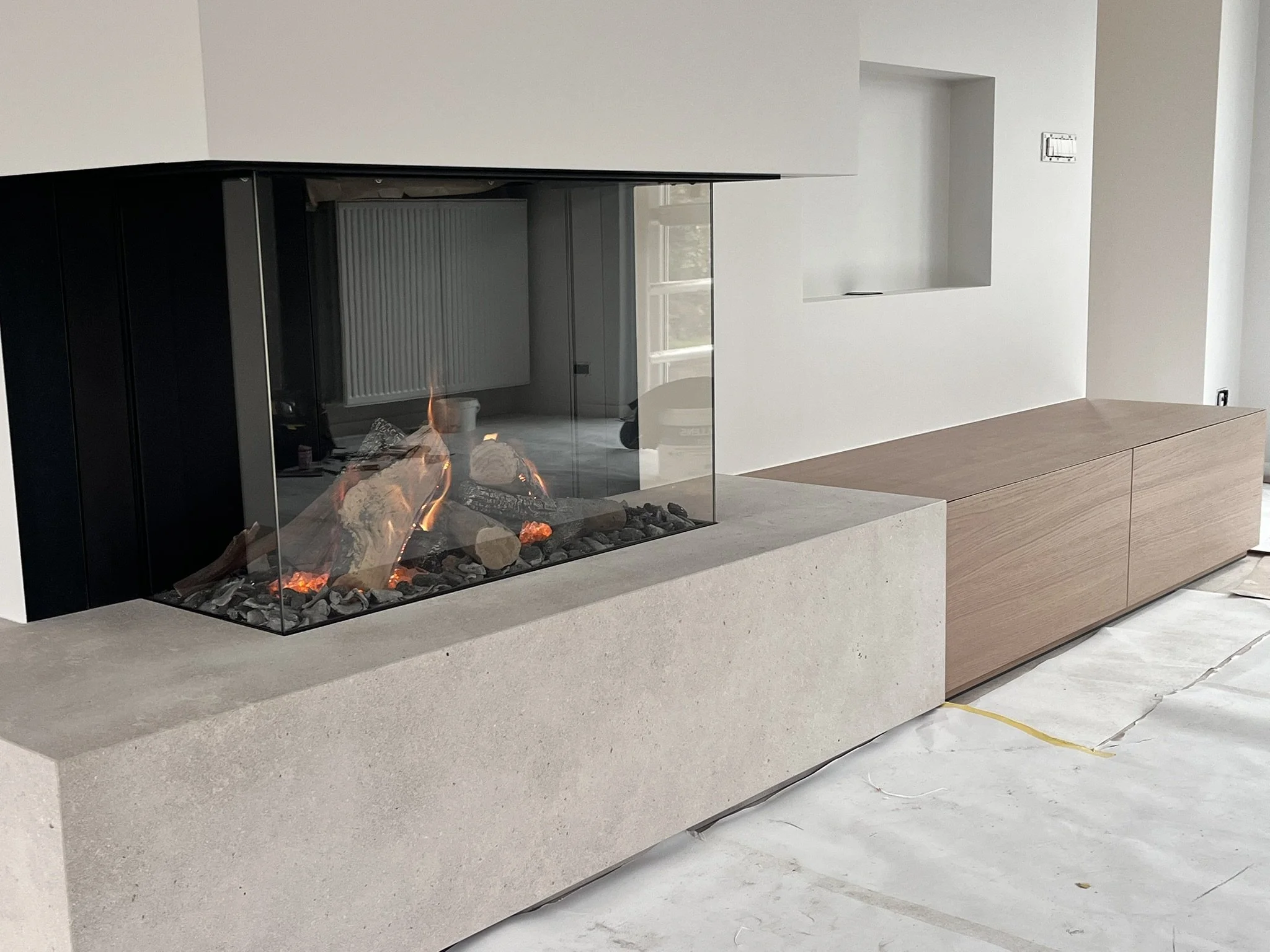 Moderne open haard met glazen omhulling en houten kasten ernaast in een witte kamer.