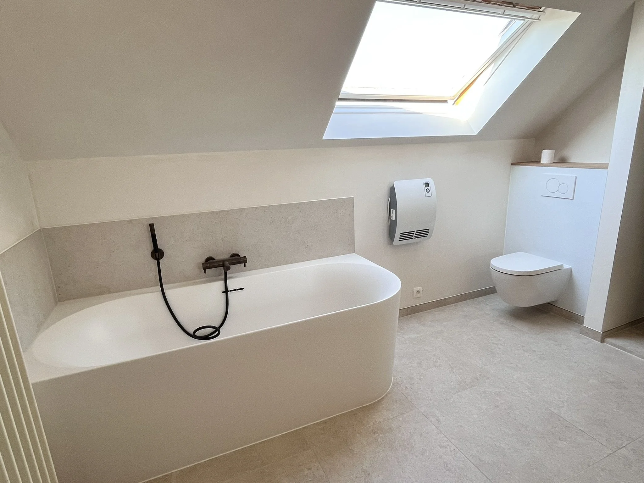 Moderne badkamer met vrijstaande badkuip onder een dakraam, toilet en handdoekradiator, lichte muren en vloer.