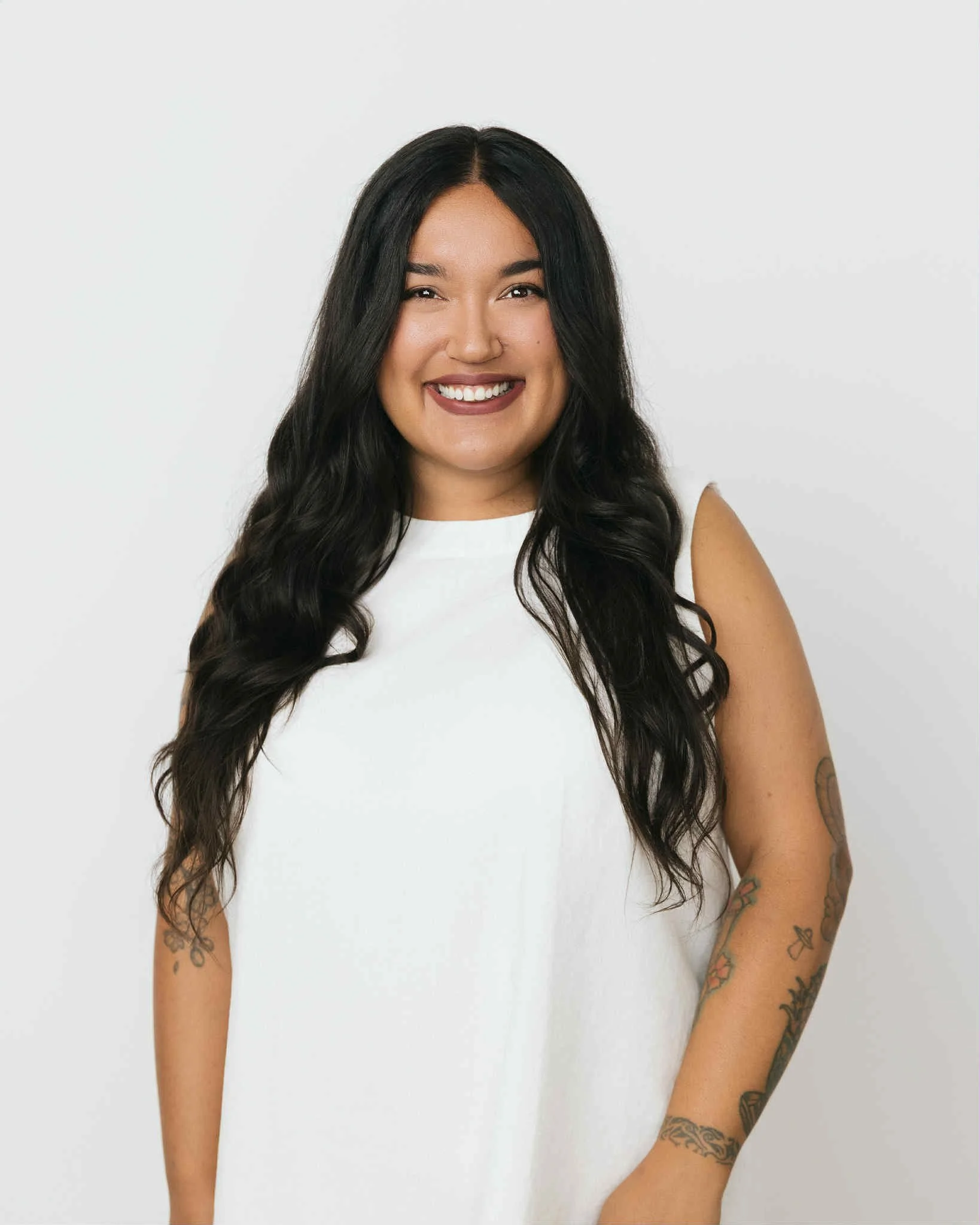 A woman with long dark wavy hair, wearing a sleeveless white top, smiling against a plain white background, with visible tattoos on her arms.