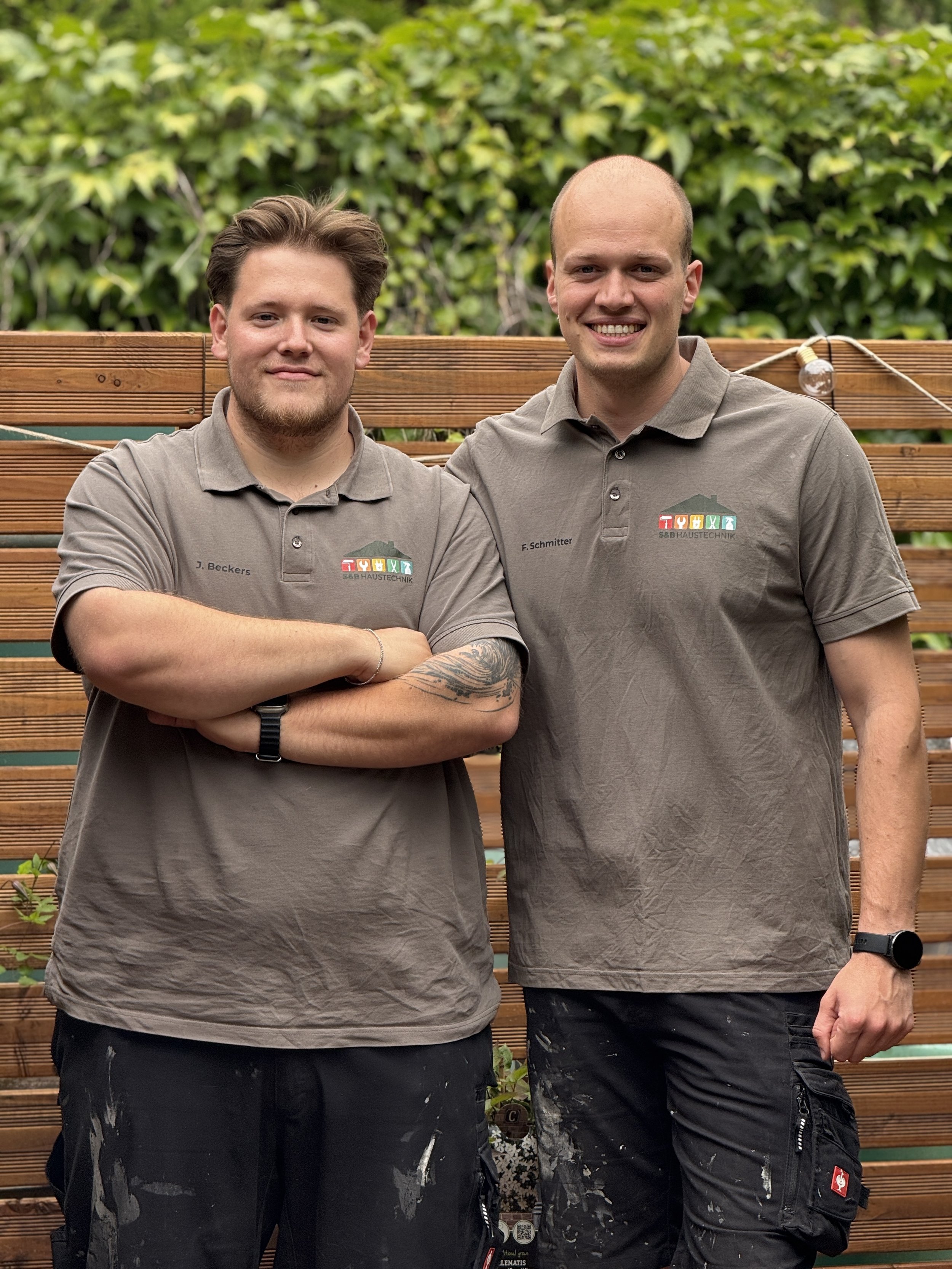 Zwei junge Männer in grauen Polohemden stehen vor einer Holzlattenwand mit grünen Blättern im Hintergrund. Beide lächeln in die Kamera, der Mann auf der linken Seite hat Arme verschränkt, der andere hat die rechte Hand an der Seite. Sie tragen schwarze Hosen mit weißer Farbe und haben Uhren an den Handgelenken.