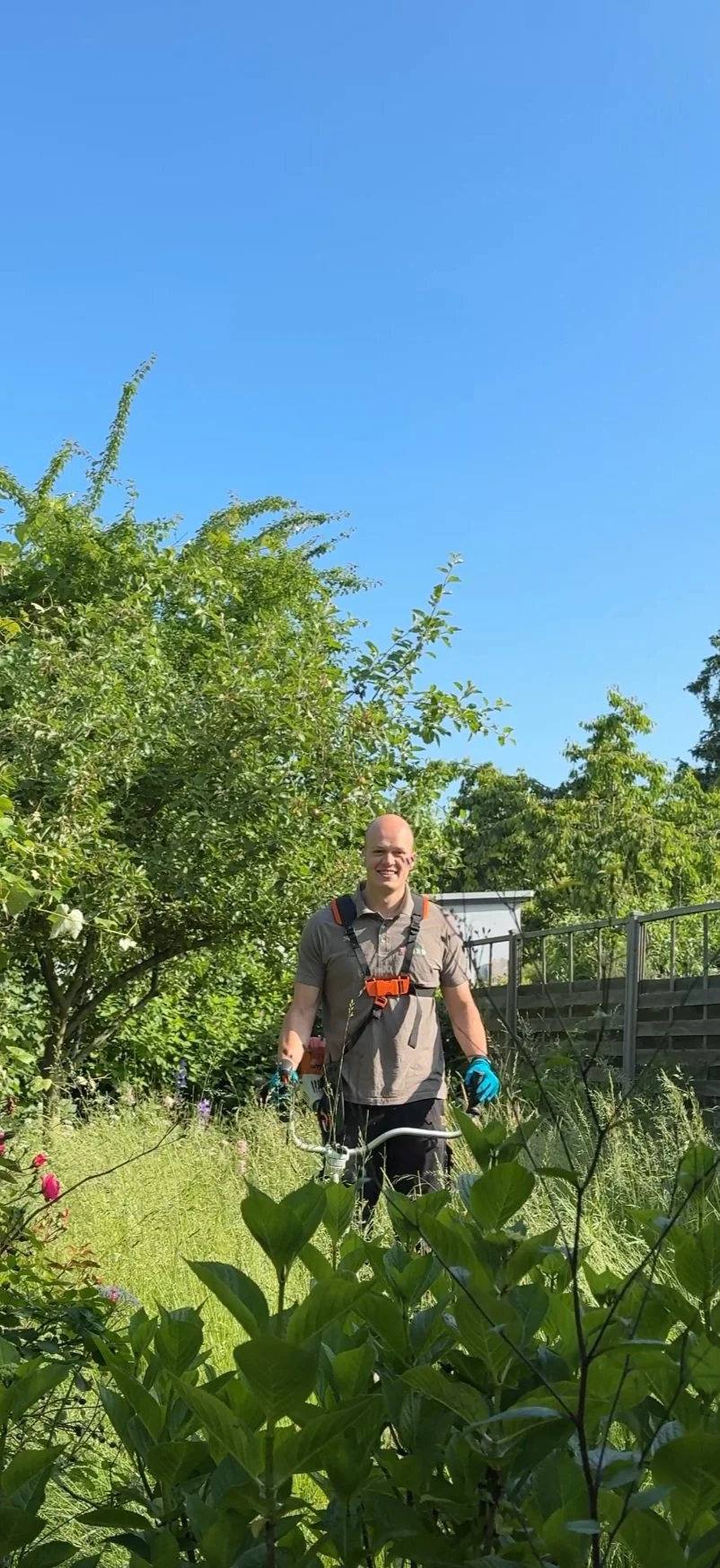 Ein lachender Mann mit Rucksack und Gartenschere steht im Garten vor grünen Büschen und Bäumen, blauer Himmel im Hintergrund.
