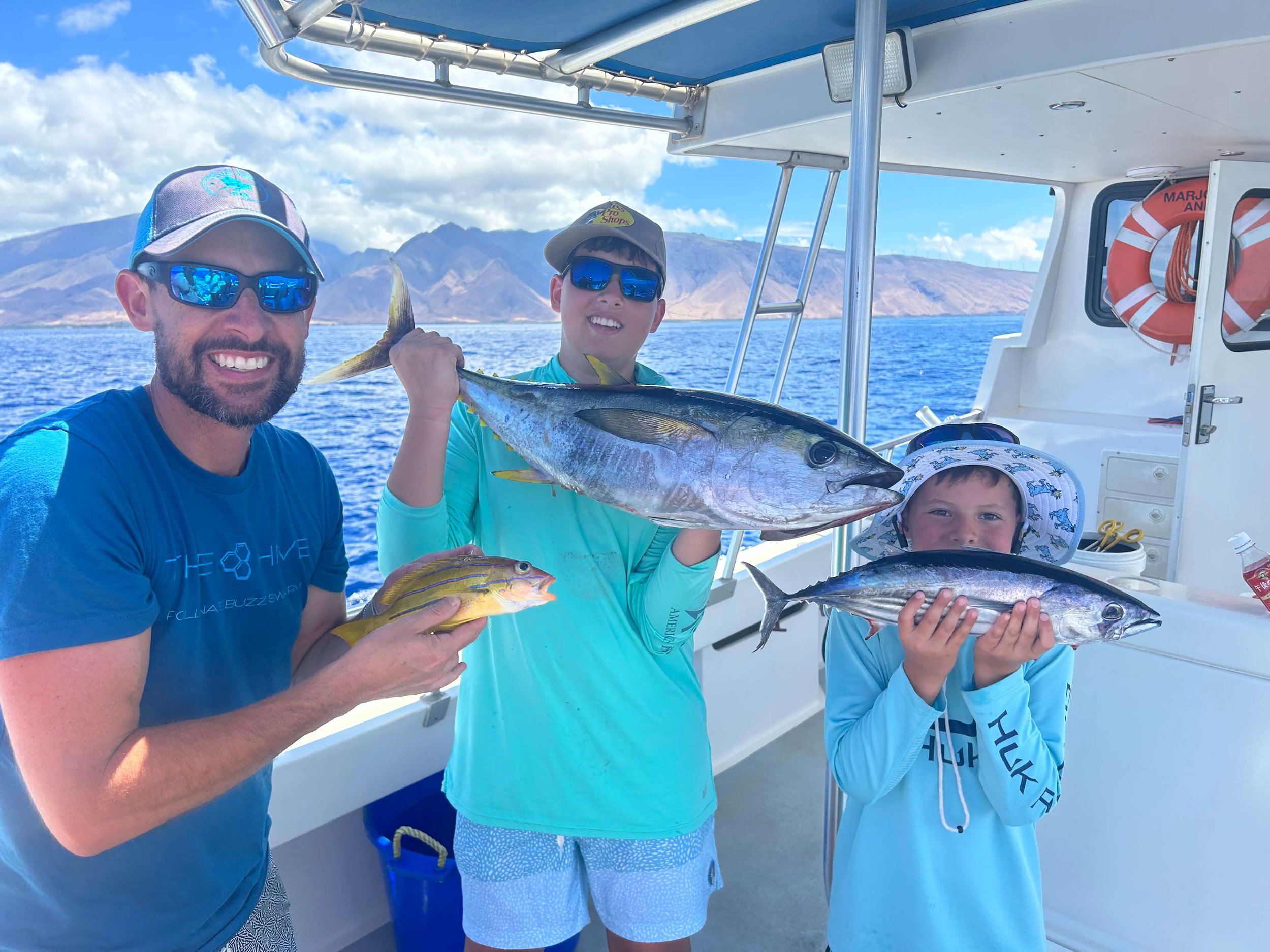 Three smiling males on a boat holding fish they caught, with a background of water and mountainous landscape.