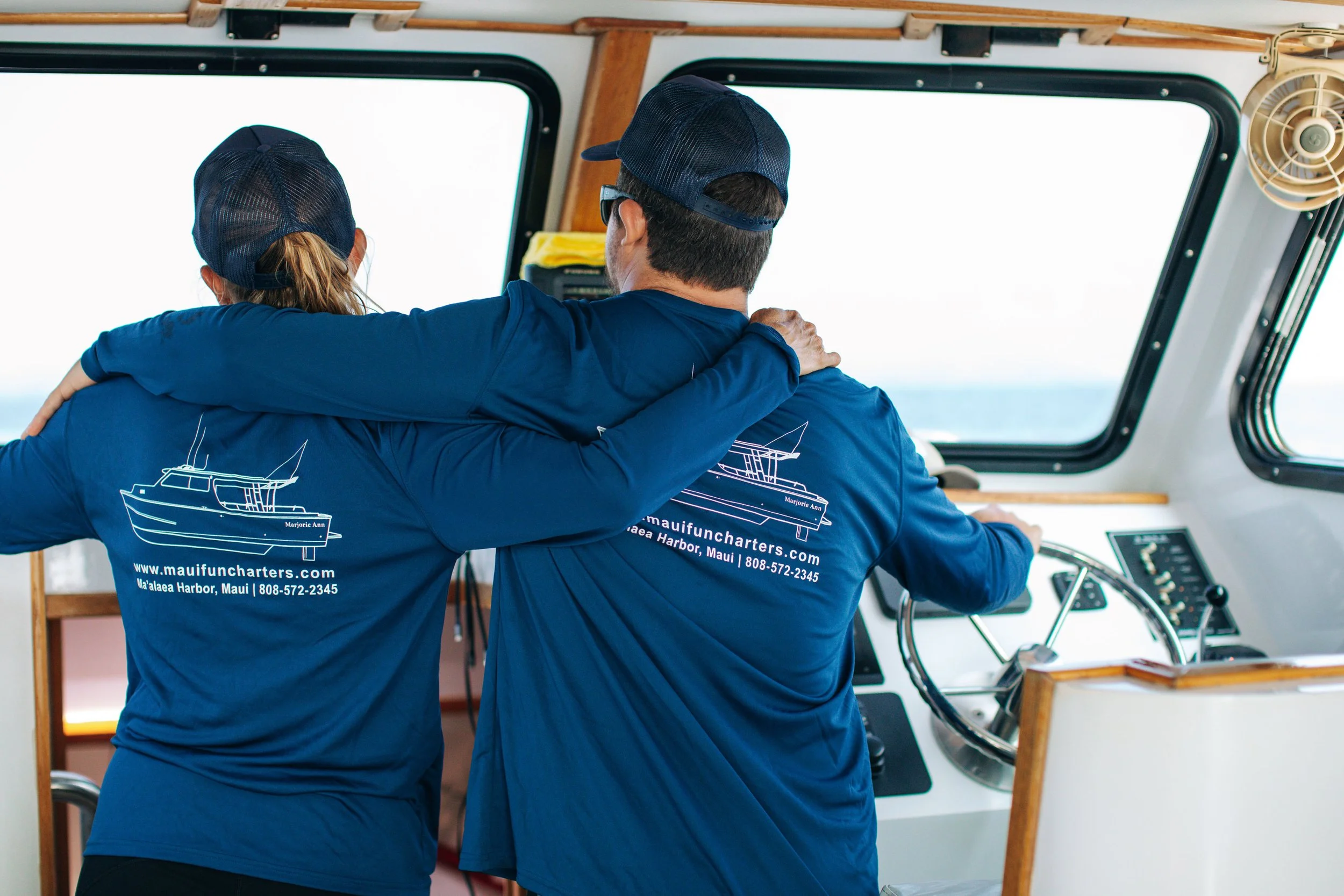 Two people in blue shirts and caps, with one person's arm around the other, on a boat navigating water.