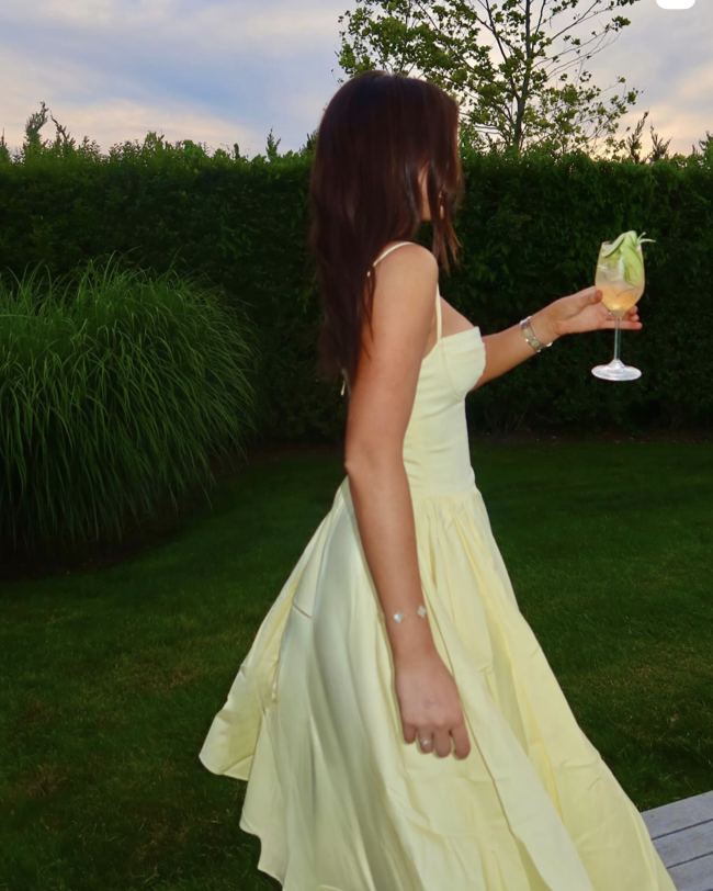 A woman in a yellow dress holding a cocktail glass with fruit and a slice of lemon, standing outdoors on a lawn with green bushes and trees in the background, during sunset.