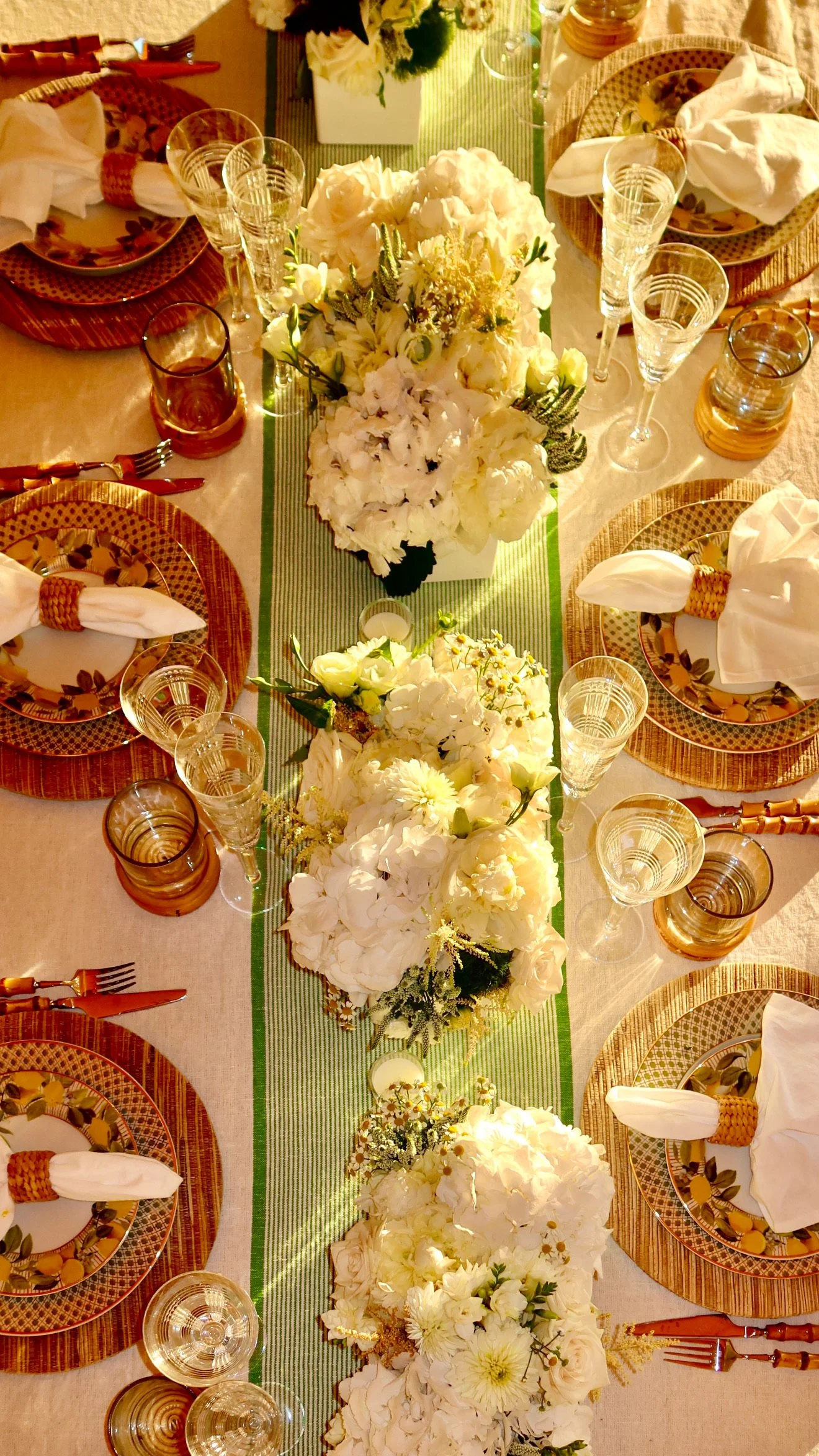 A top view of a table set for a meal with white floral centerpieces, wine glasses, plates with napkins, and cutlery. The table has a white tablecloth with a green striped table runner.