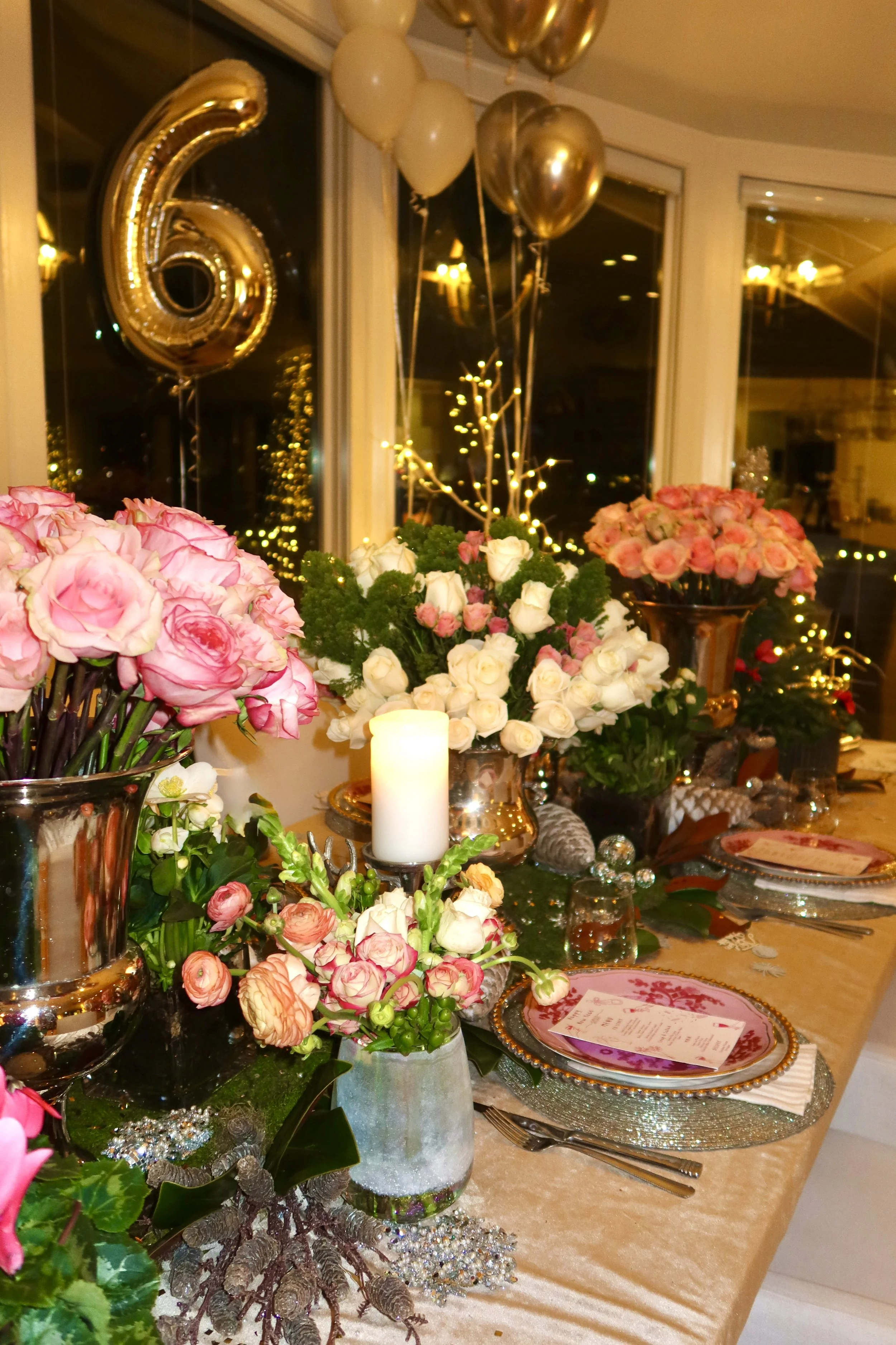 A table decorated for a 60th birthday celebration with flowers, balloons, a candle, and dinnerware, viewed near a window at night.