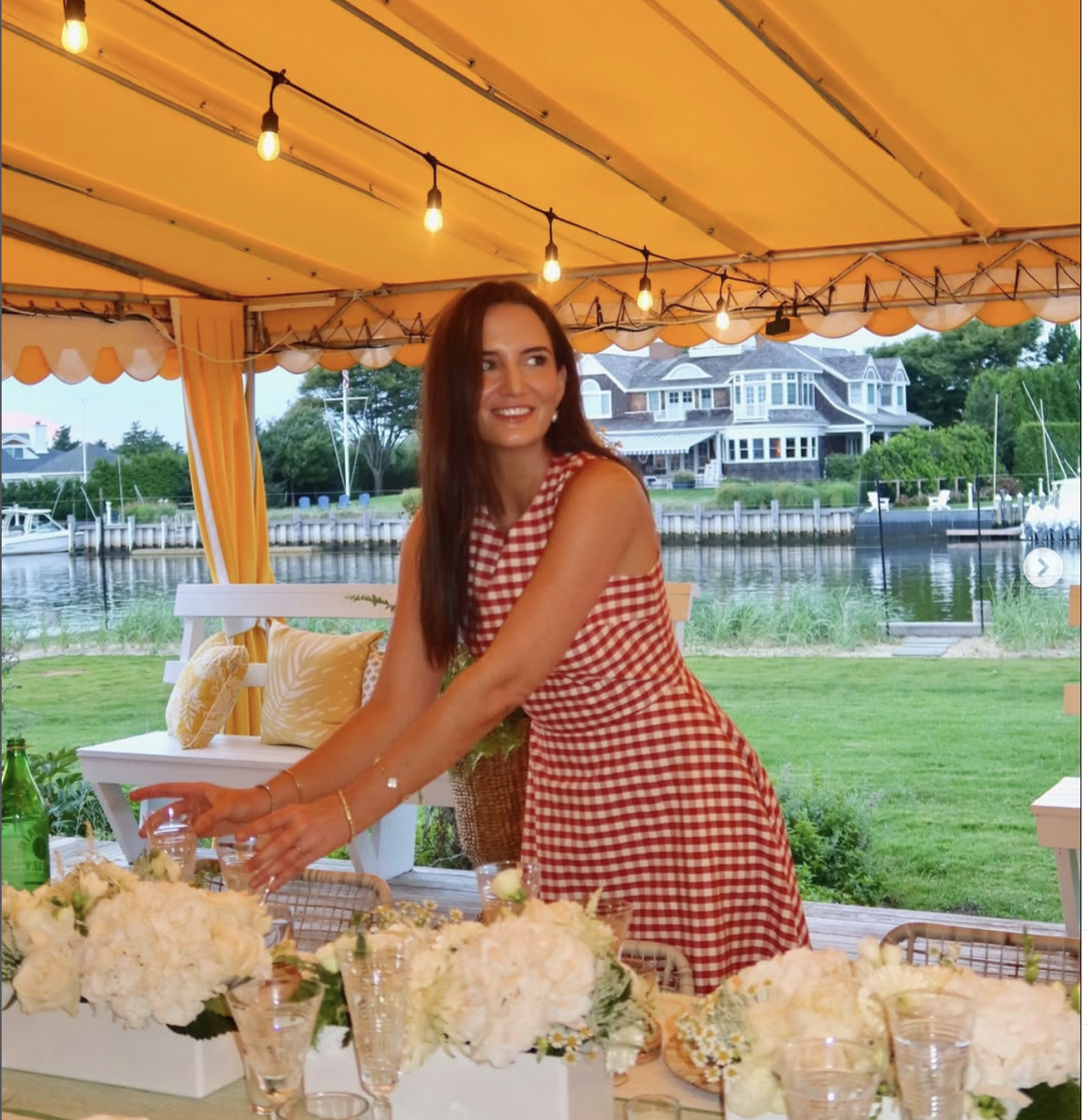 Woman in a red and white checkered dress at an outdoor event, surrounded by floral arrangements, with a house and water in the background.