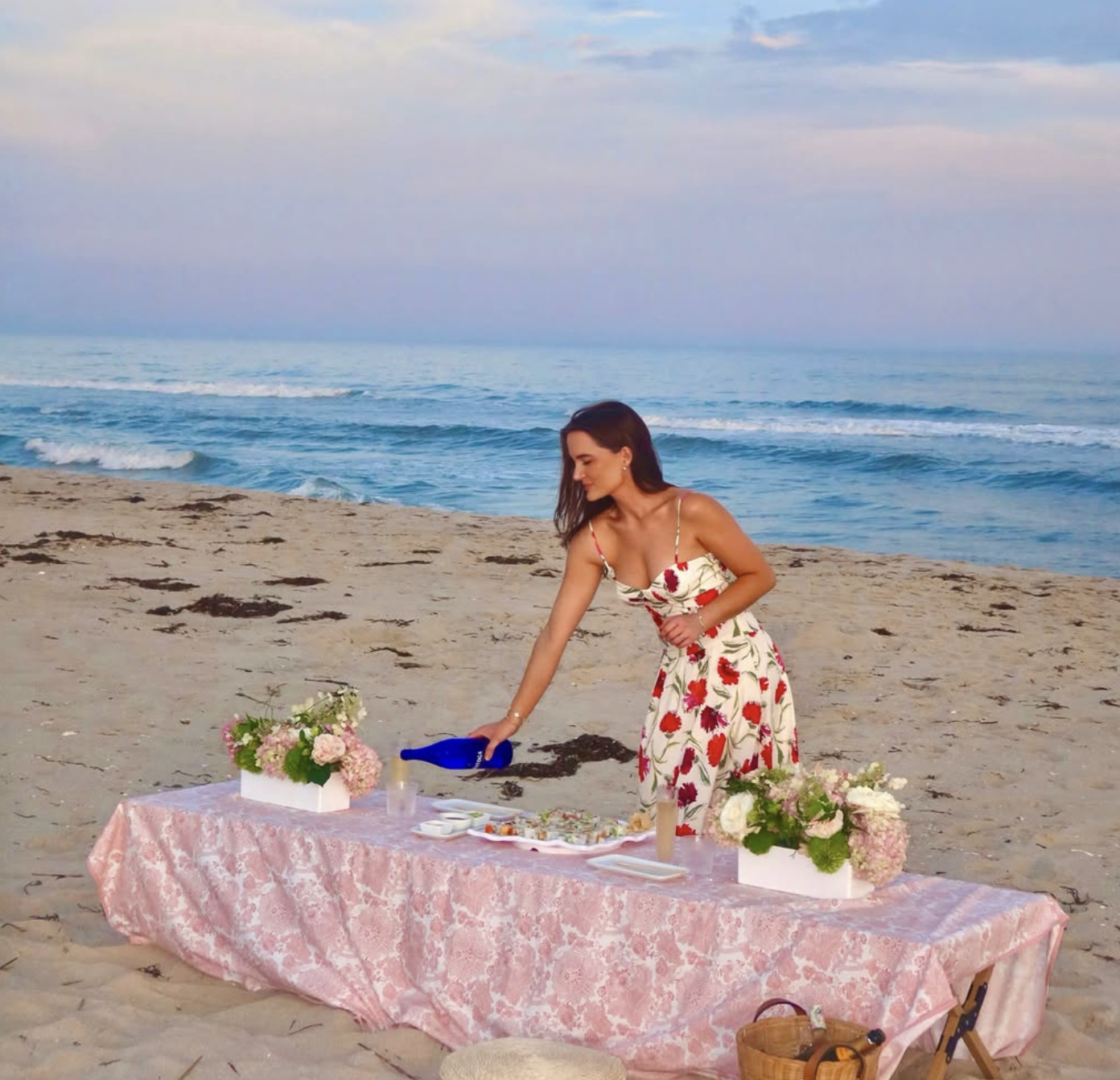 A woman in a floral dress arranging flowers on a table set up on the beach with the ocean in the background at sunset.