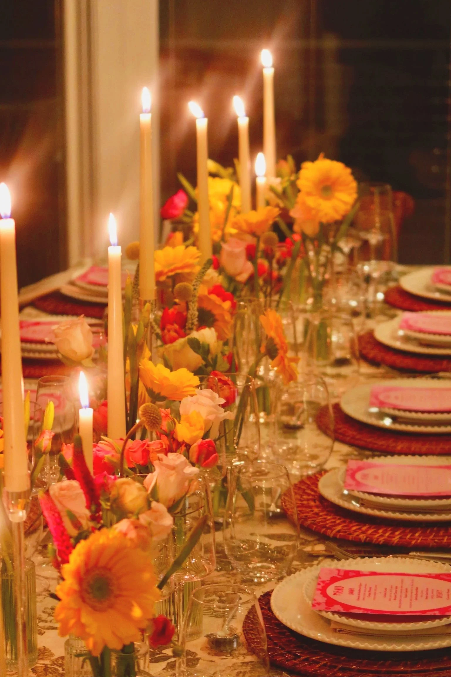 A dinner table decorated with colorful flowers, tall lit candles, and place settings for a meal.
