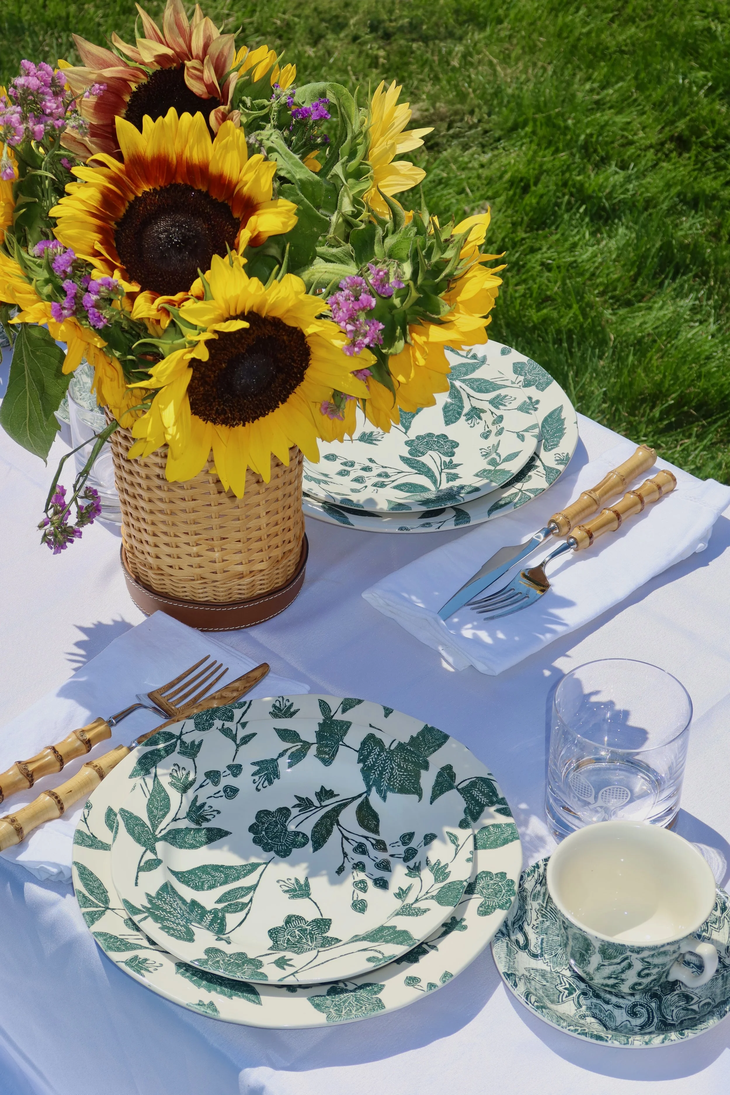 A round table set outdoors with a sunflower and purple flower bouquet in a wicker basket, green patterned dishes, gold-handled cutlery, a glass of water, and a cup of cream or milk, on a white tablecloth with grass in the background.