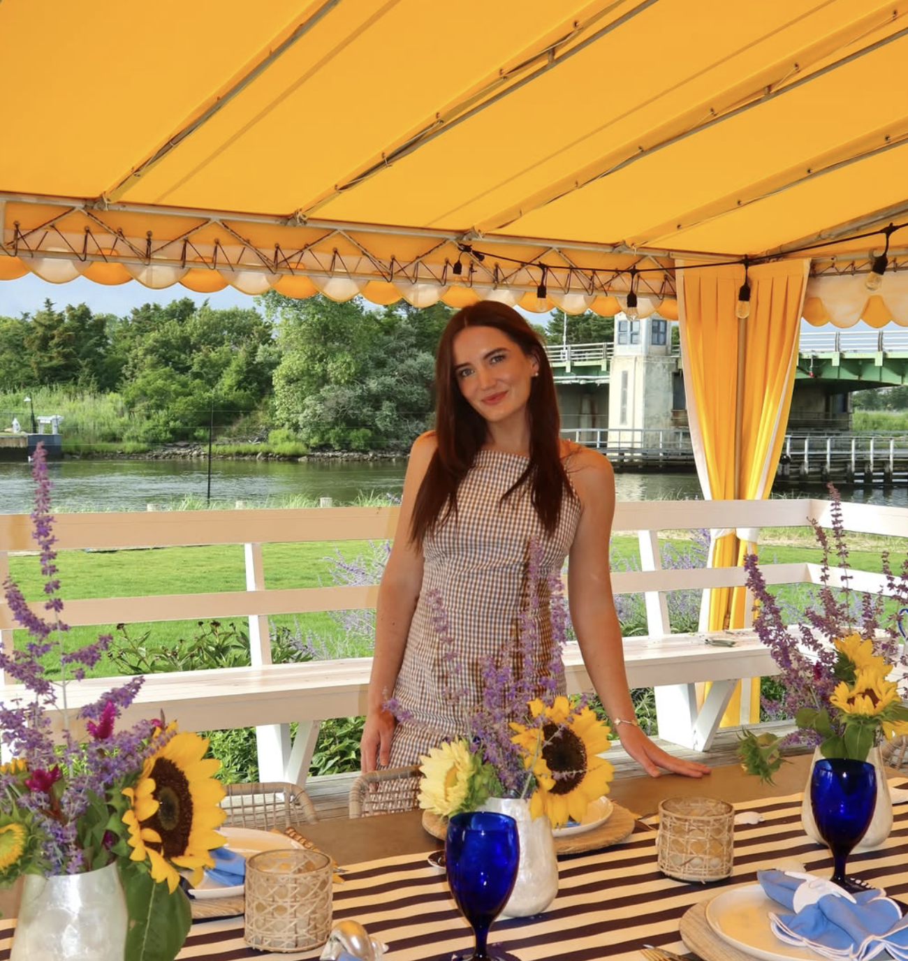 A woman with long brown hair smiling at a dining table decorated with sunflowers and purple flowers, with a river and lush green trees in the background, under a yellow canopy with curtains.