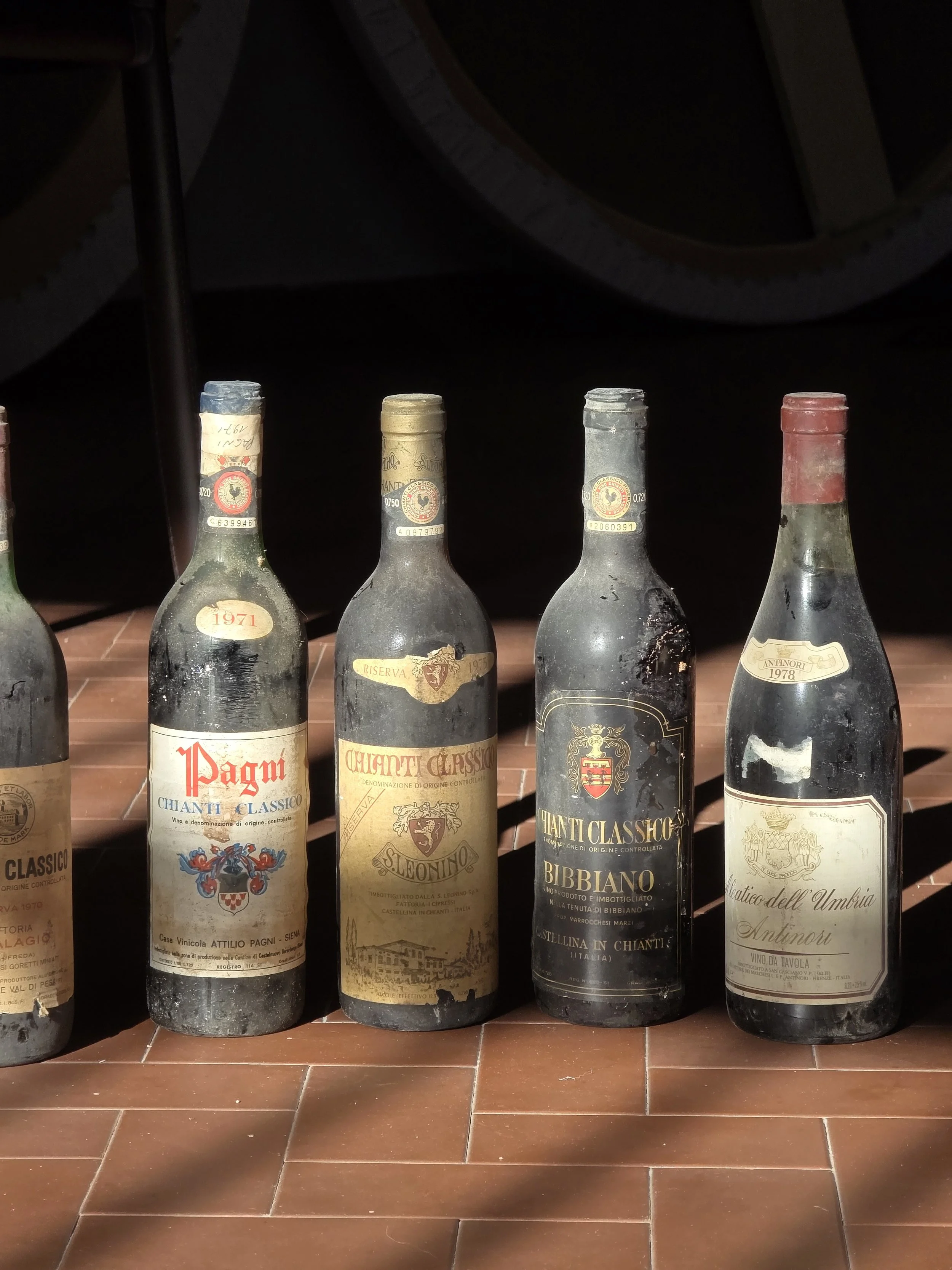 Quattro bottiglie di vino vecchie e polverose su una superficie di piastrelle di colore marrone, con uno sfondo scuro.