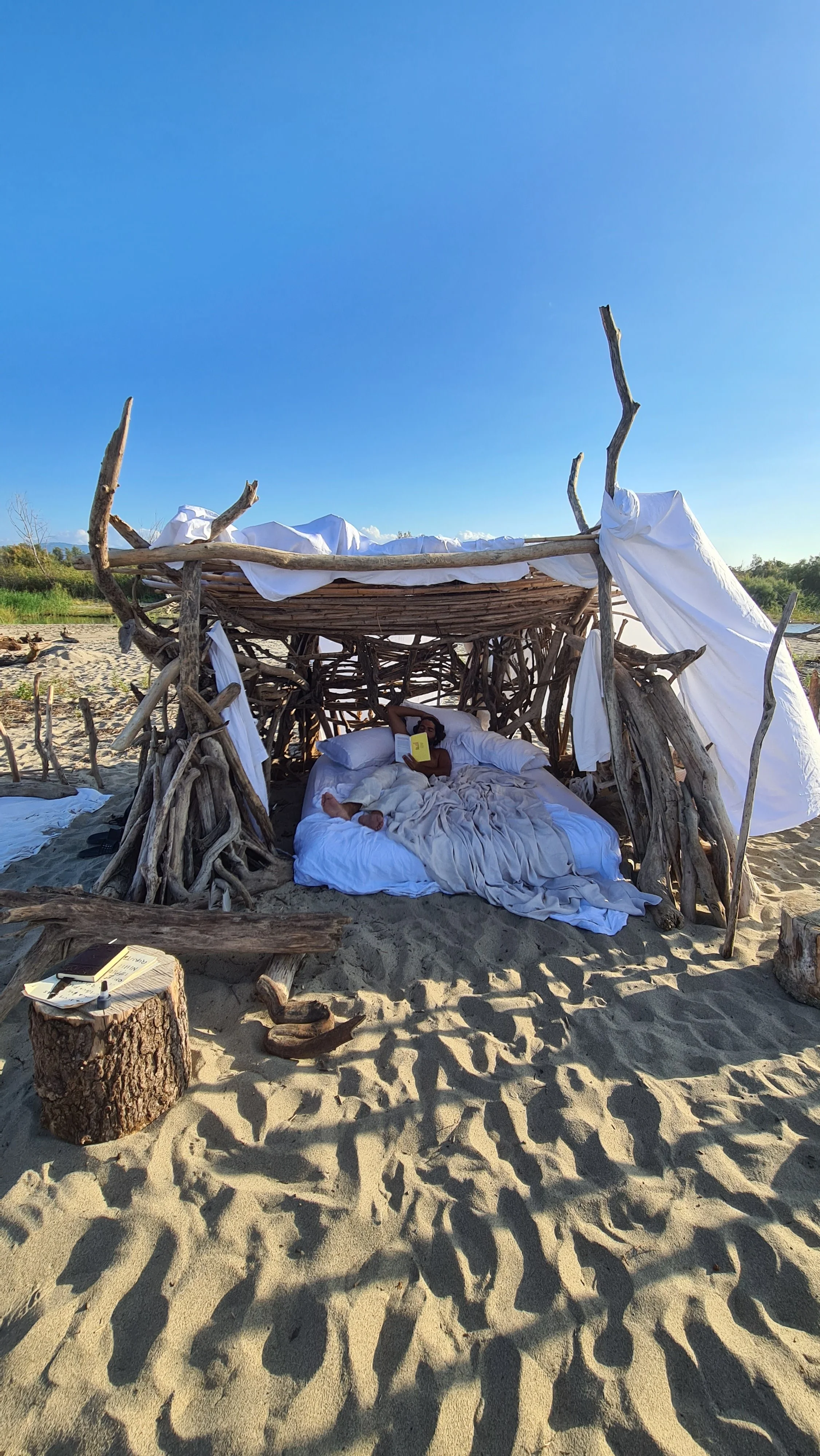 Struttura di legno sulla spiaggia con un tetto di rami e teli bianchi, una persona (ROMANO OLIVERI) sdraiata sul letto a leggere un libro, con un tavolo di tronco e un block notes accanto. ARNO STUDIO