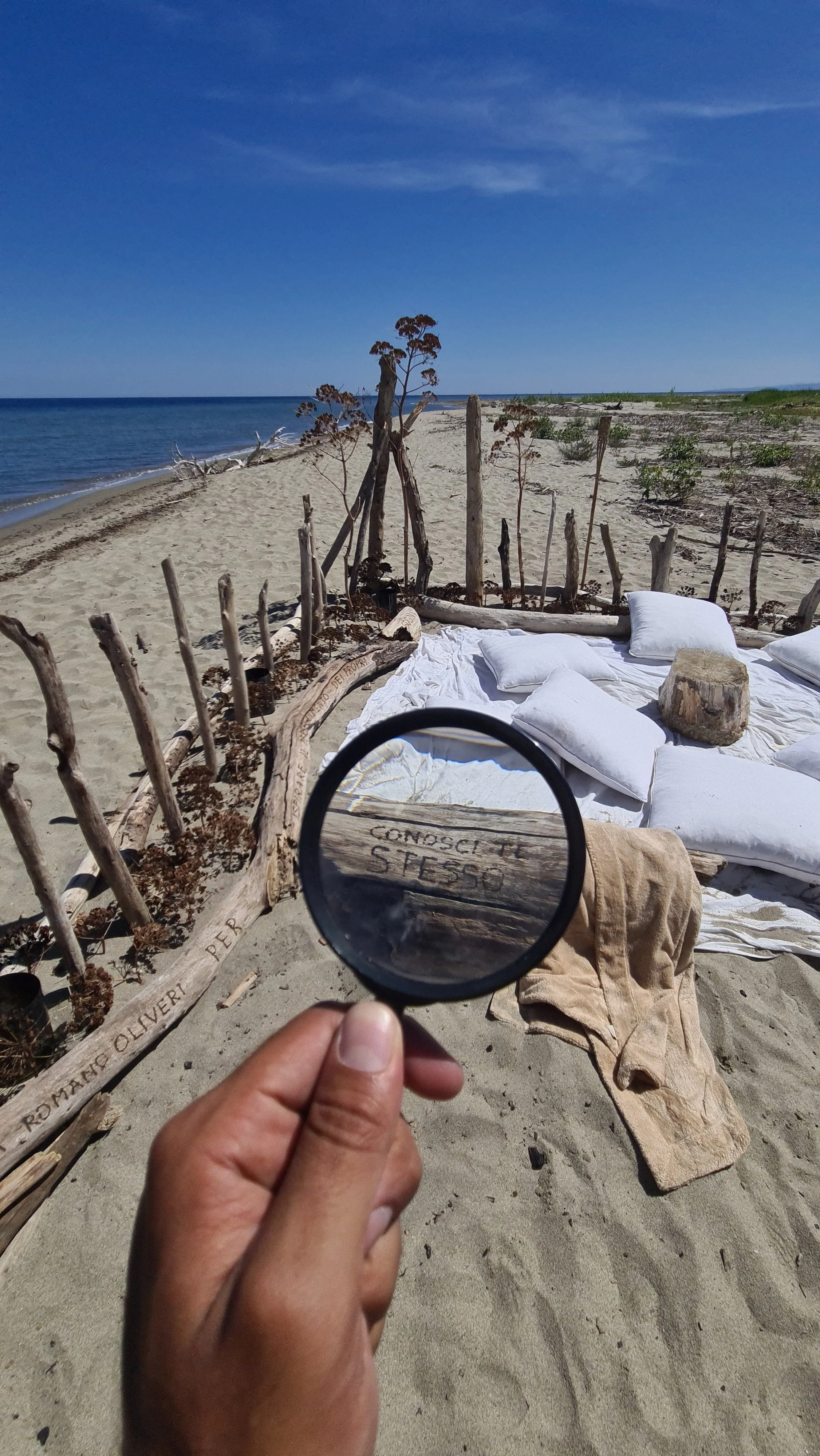 Spiaggia con tende fatte di legno e tessuto bianco, alcune coperte e cuscini, vista del mare e cielo azzurro, con una mano che tiene una lente d'ingrandimento che evidenzia una scritta sul legno."CONOSCI TE STESSO".