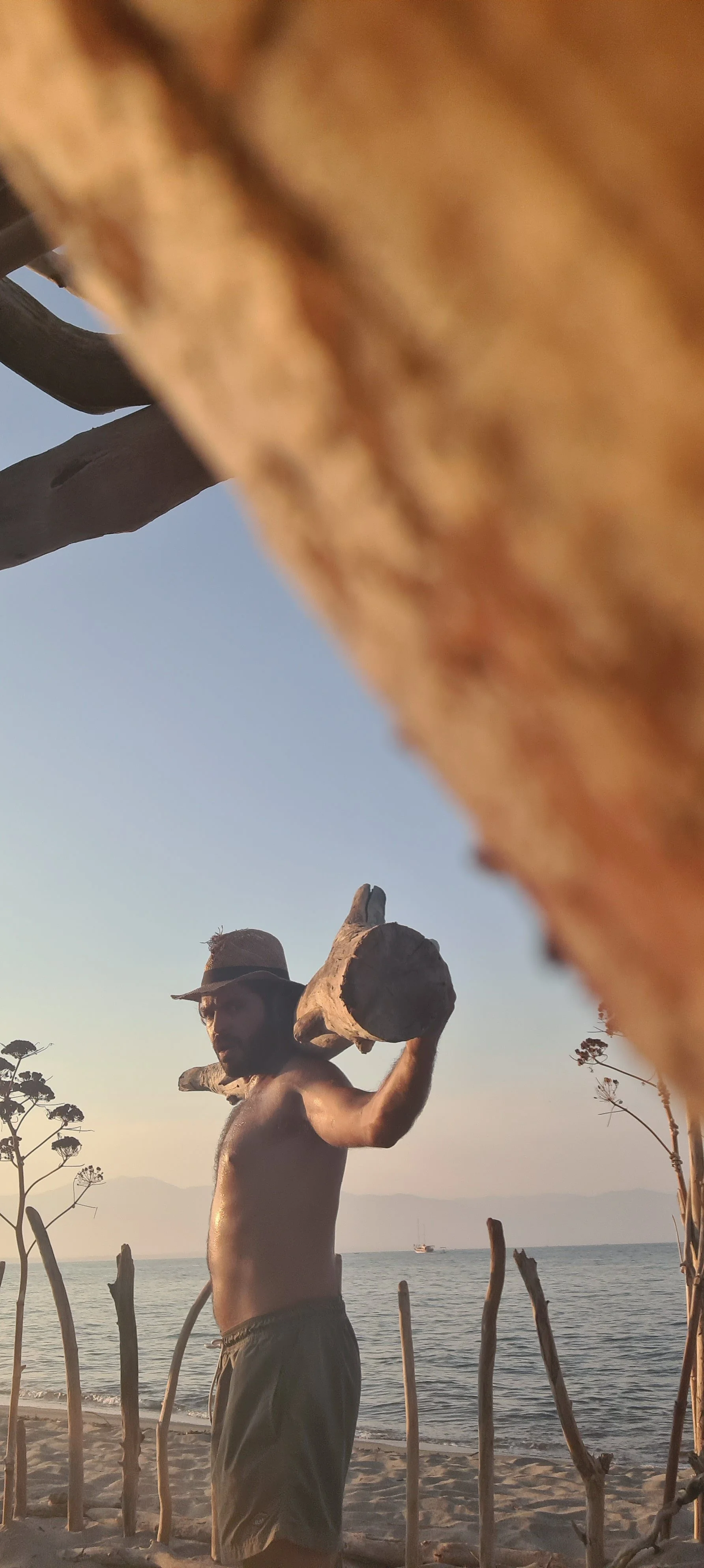 Uomo senza maglietta con cappello, che tiene un grande pez di legno sulla spalla sulla spiaggia, con il mare e le montagne sullo sfondo