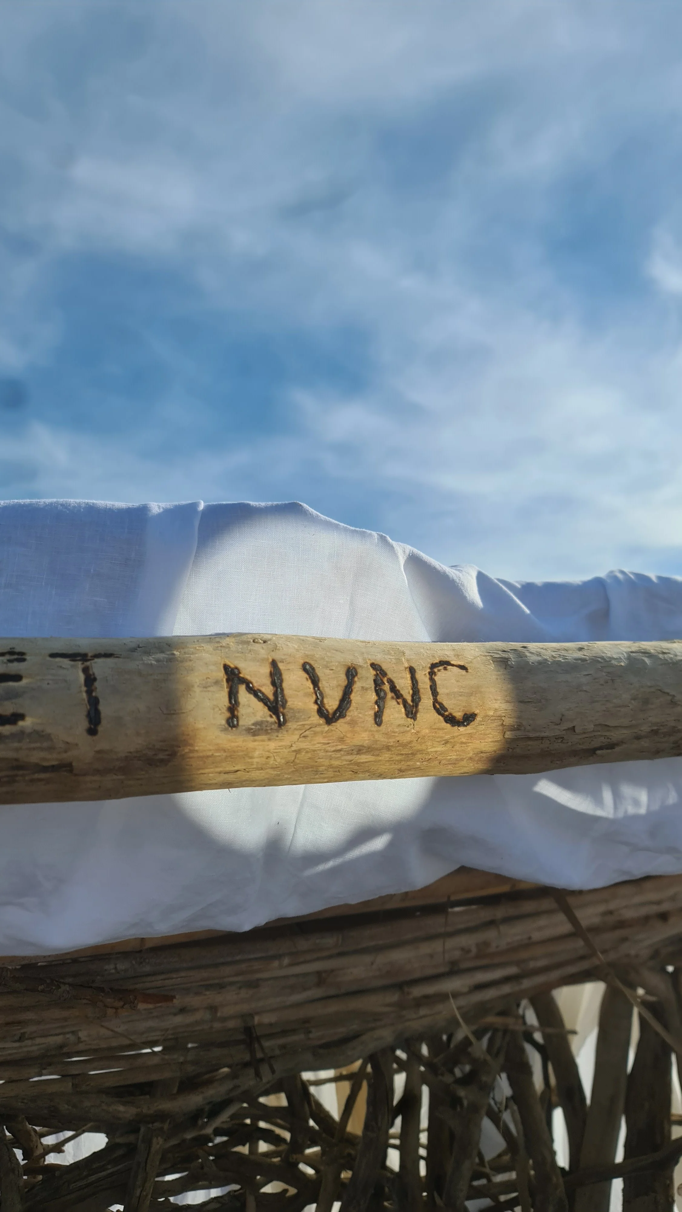 Ramo di legno con scritto 'NUNC' e tessuto bianco sotto un cielo nuvoloso.