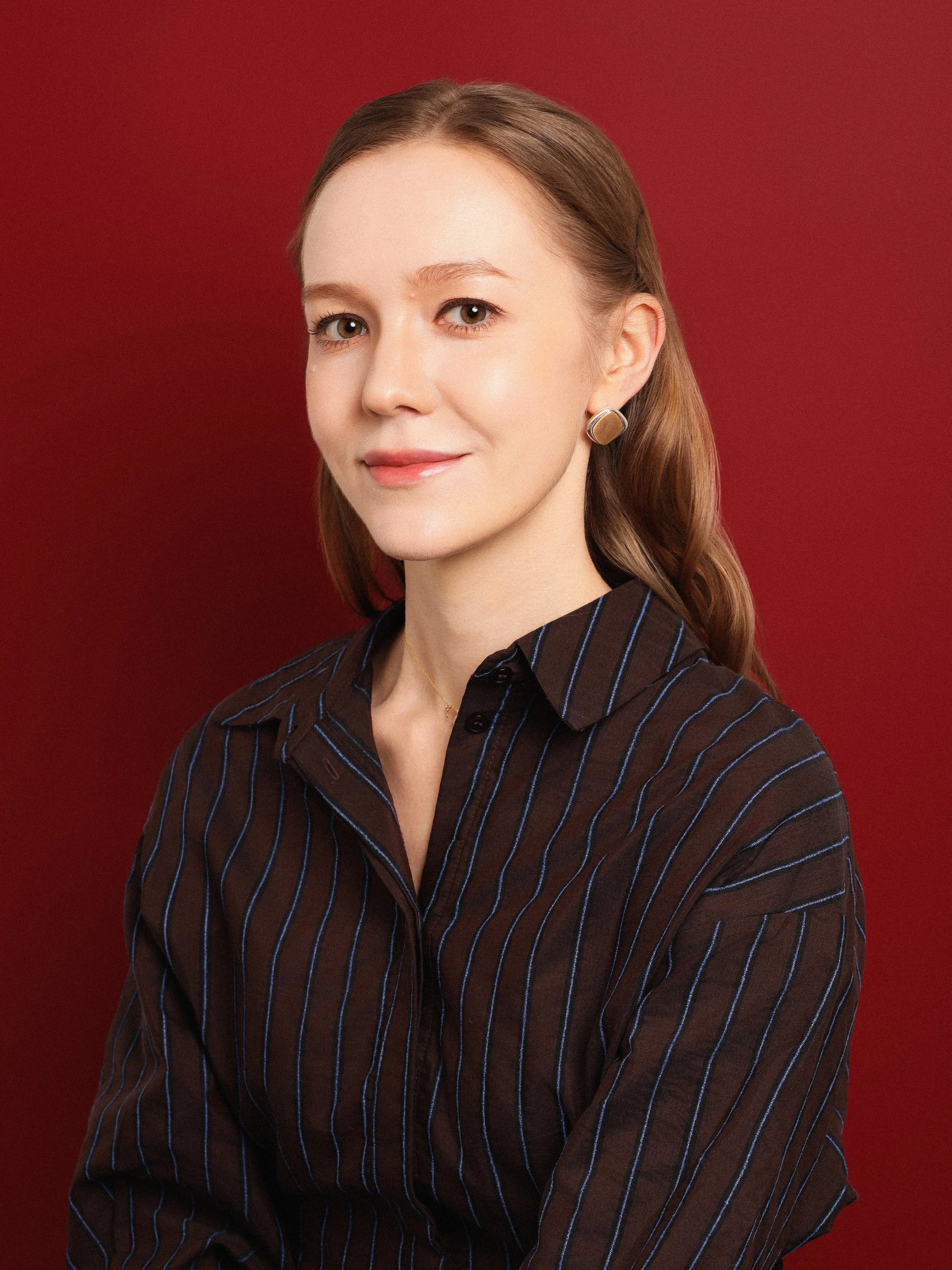 A woman with fair skin and light brown hair styled in loose waves. She is wearing a dark, striped shirt and metallic round earrings, and she is smiling softly against a dark red background.