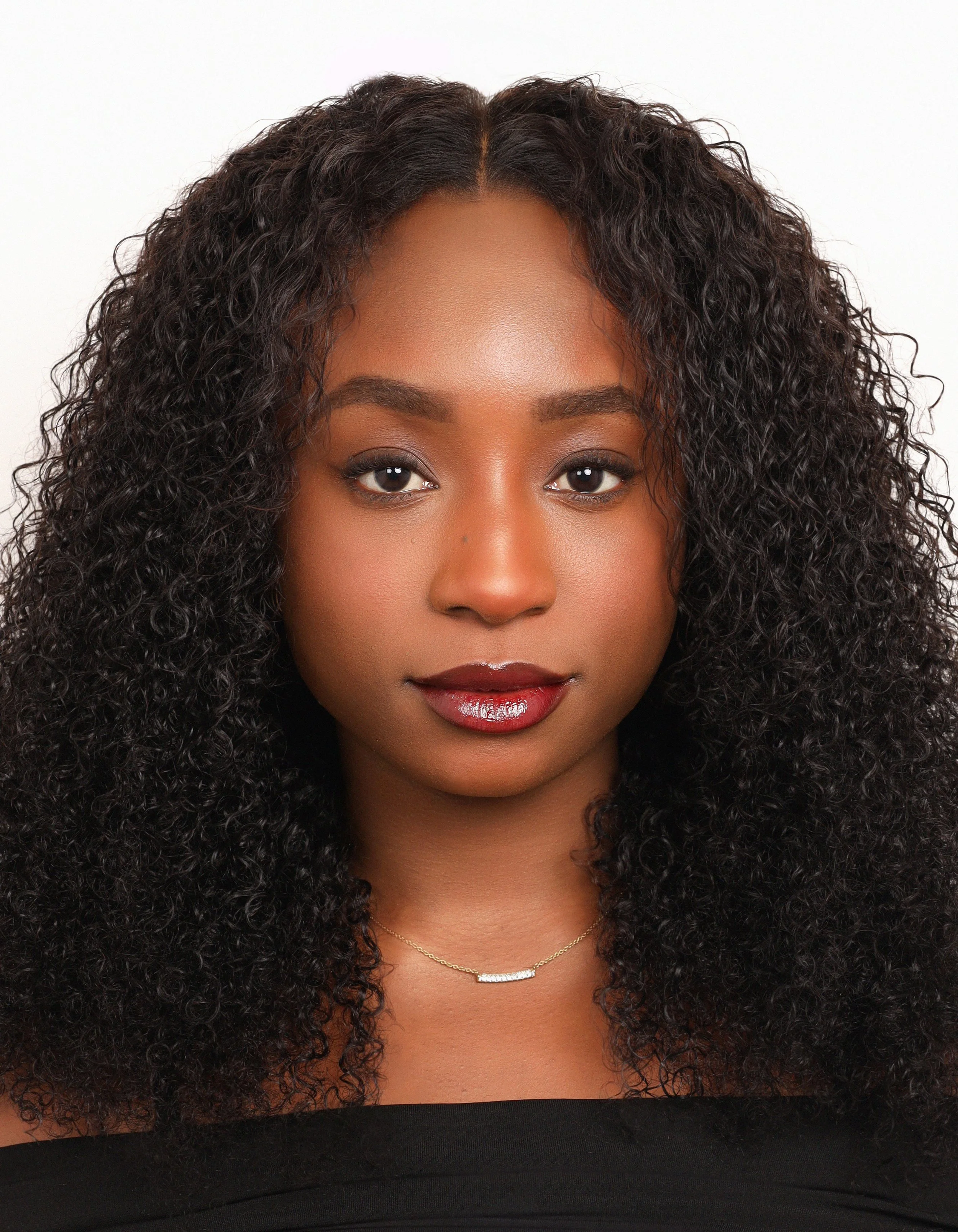 A young woman with curly black hair and brown skin looking directly at the camera, wearing a black top and a delicate gold necklace with a small bar pendant.