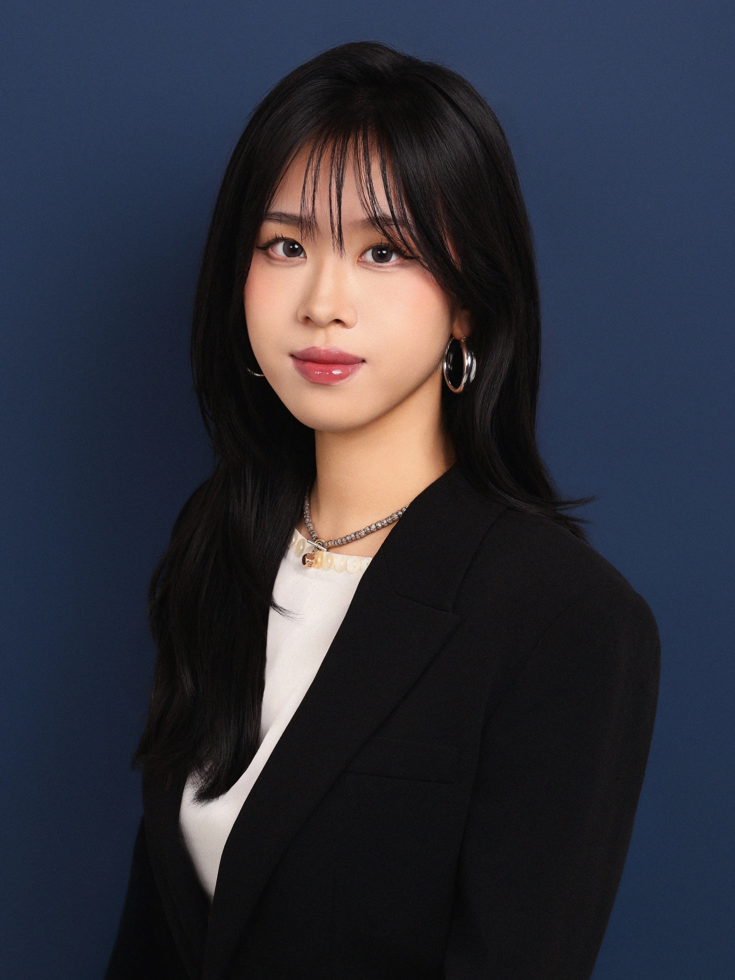 A woman with long black hair, wearing hoop earrings, a necklace, and a black blazer, posing against a dark blue background.