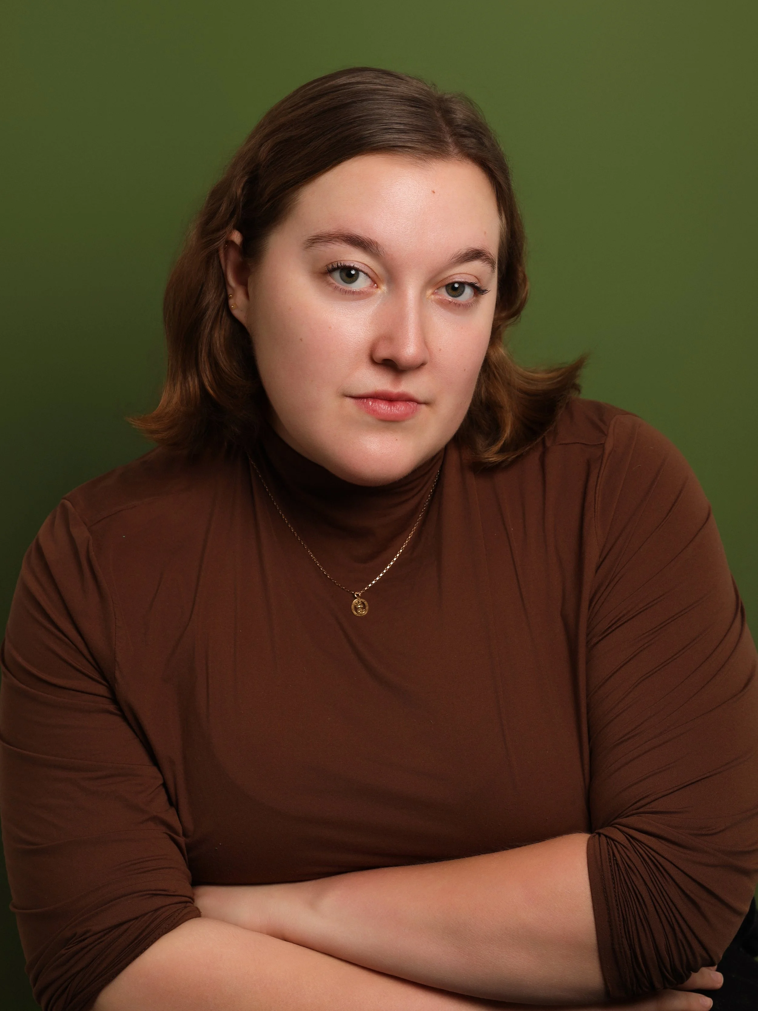 A young woman with shoulder-length brown hair, wearing a brown turtleneck and gold necklace, posing with arms crossed against a green background.