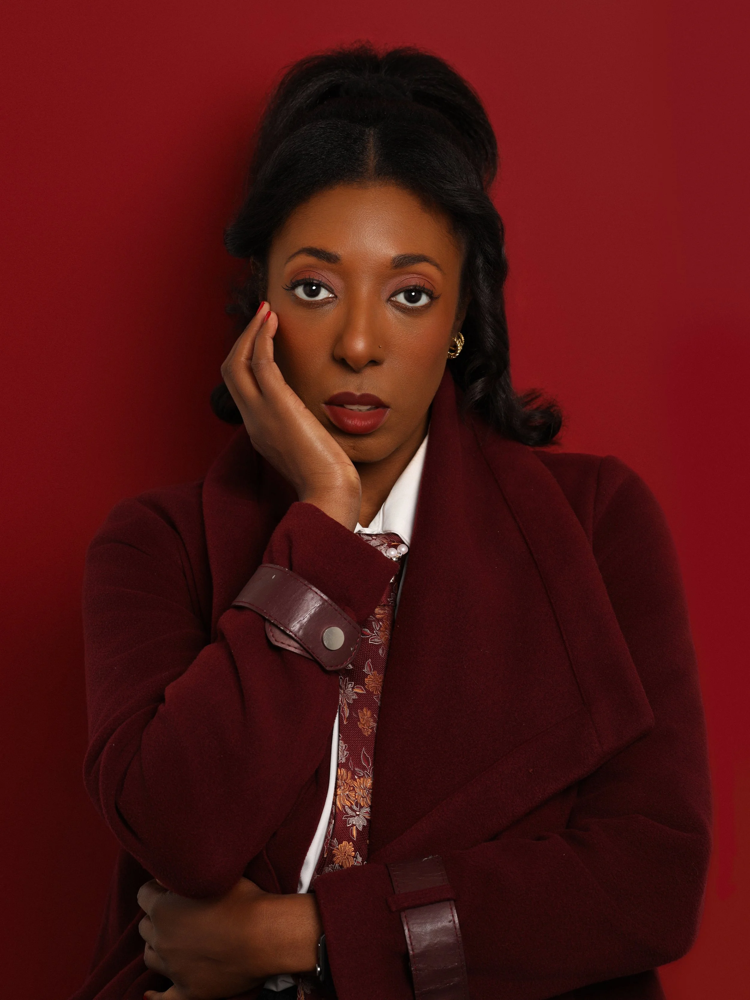 A woman with dark hair wearing a red blazer, a floral shirt, and jewelry, posing with one hand on her face against a red background.