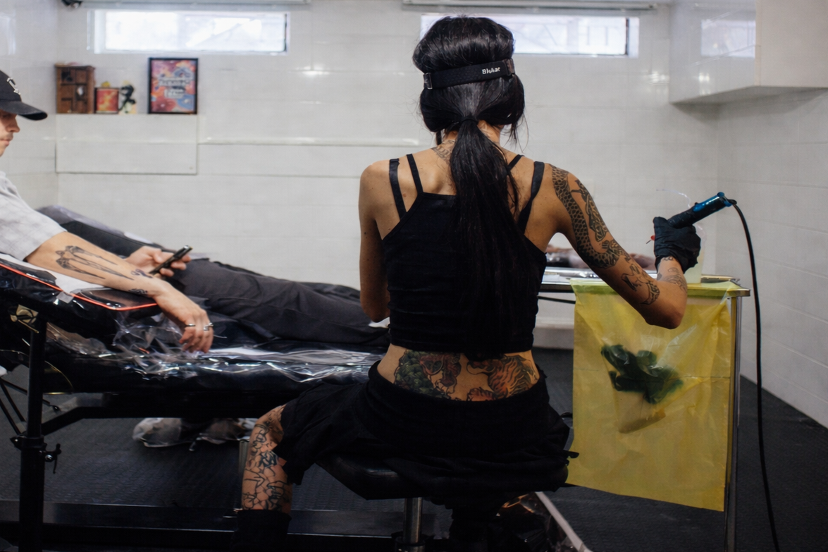 Tattoo artist with tattoos on arms and abdomen, wearing a black tank top and gloves, working on a tattoo in a studio with a client lying on a chair.