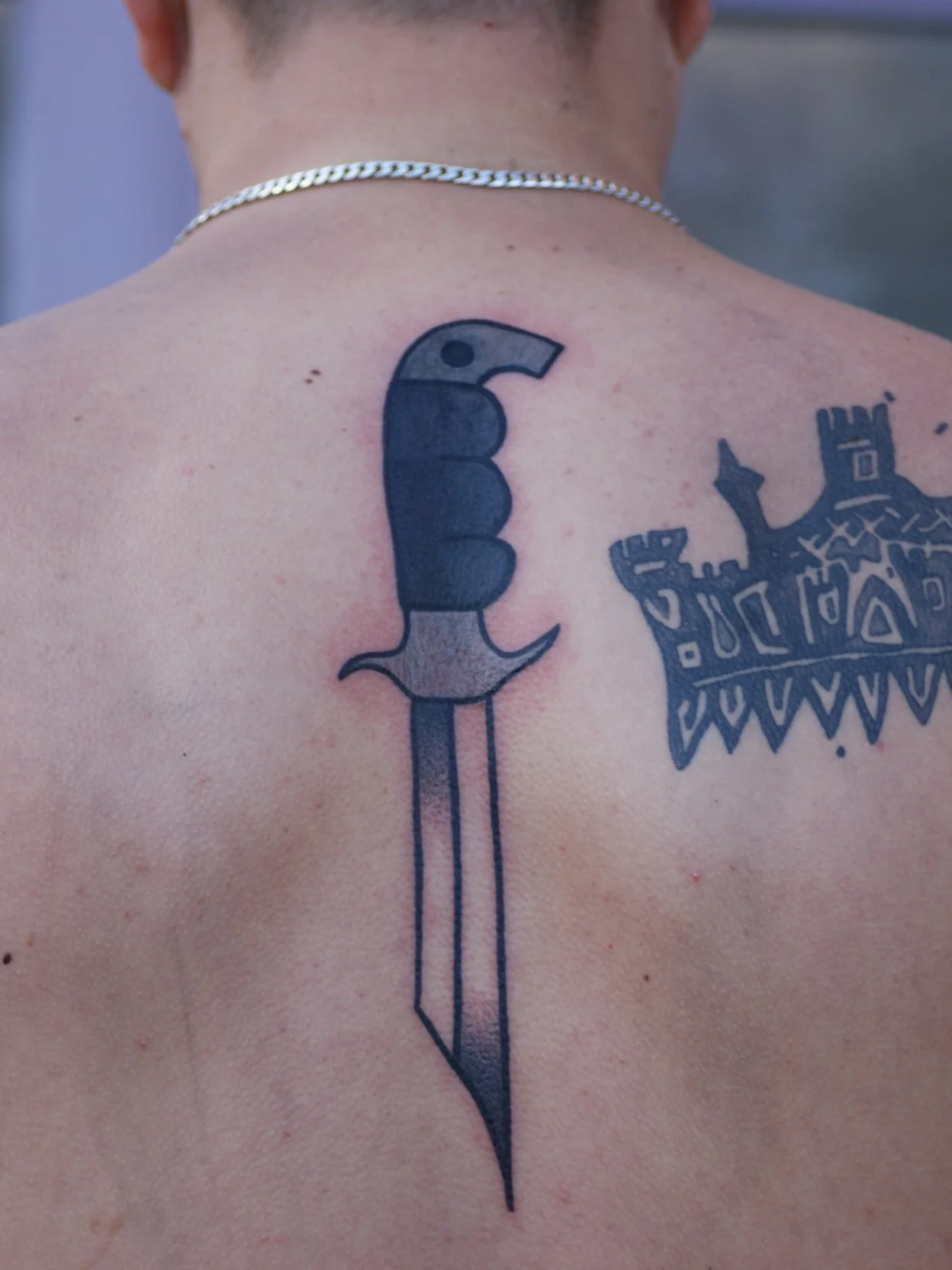 A person with tattoos on their upper back, including a black knife tattoo and a tribal-style pattern.
