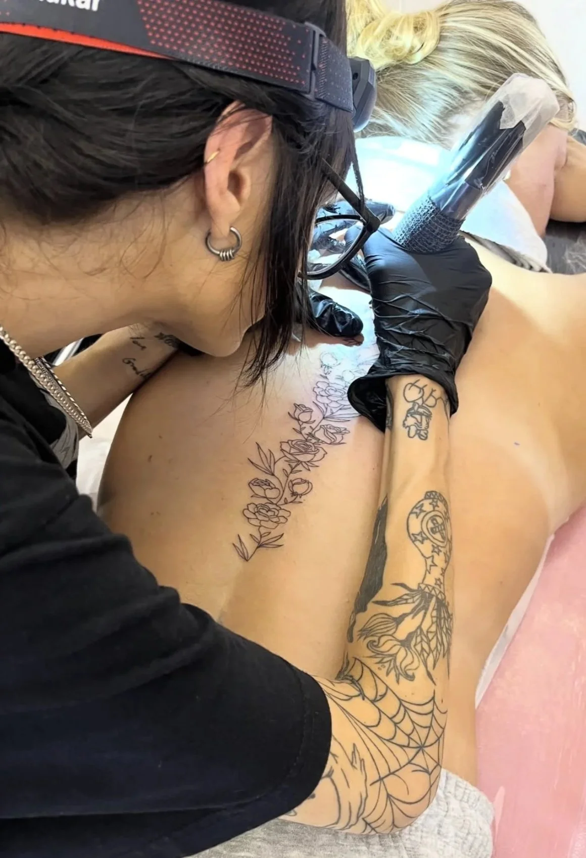 Tattoo artist in black gloves and glasses working on a person's spine tattoo, which features flowers and botanicals.