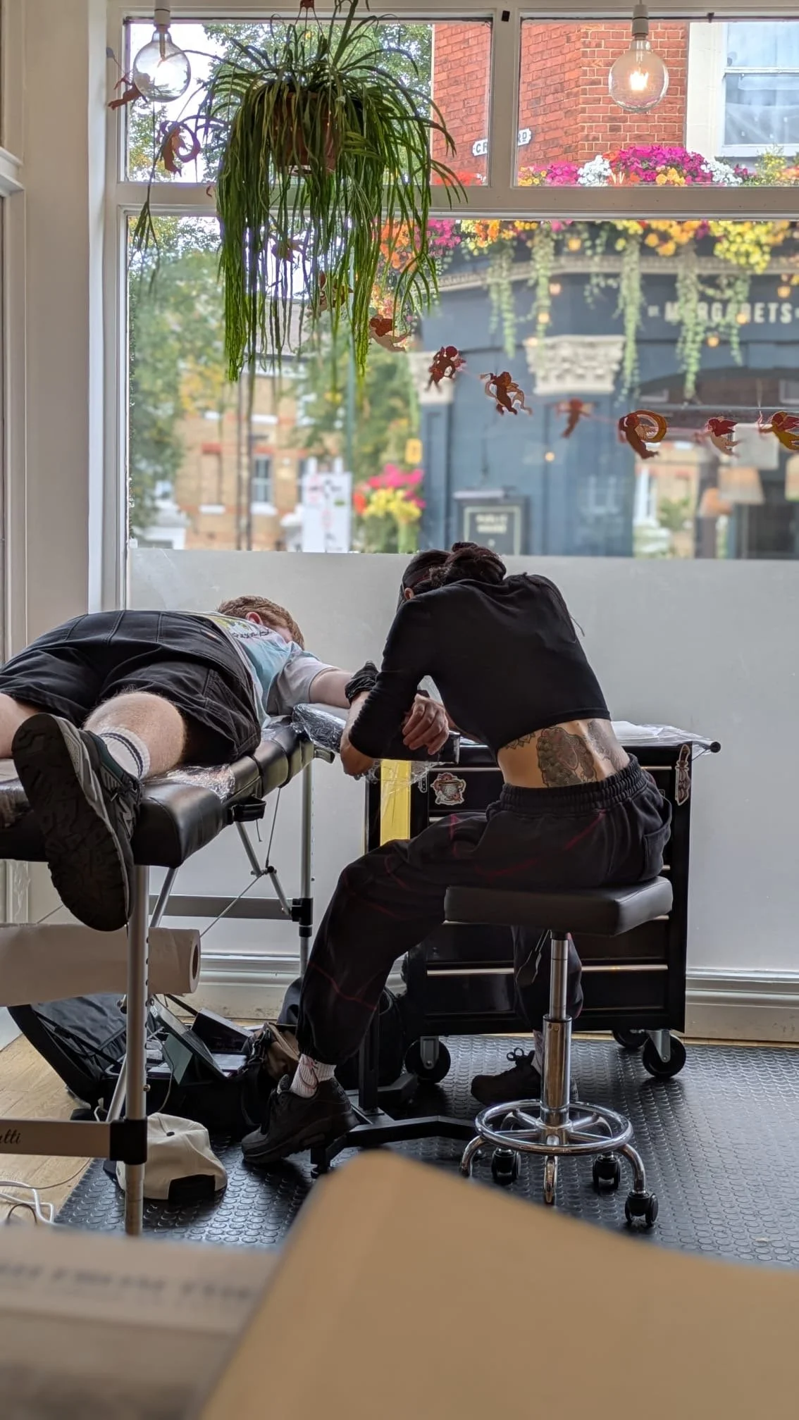 A person with tattoos on their side getting a tattoo from a tattoo artist inside a shop with large window and hanging plants.