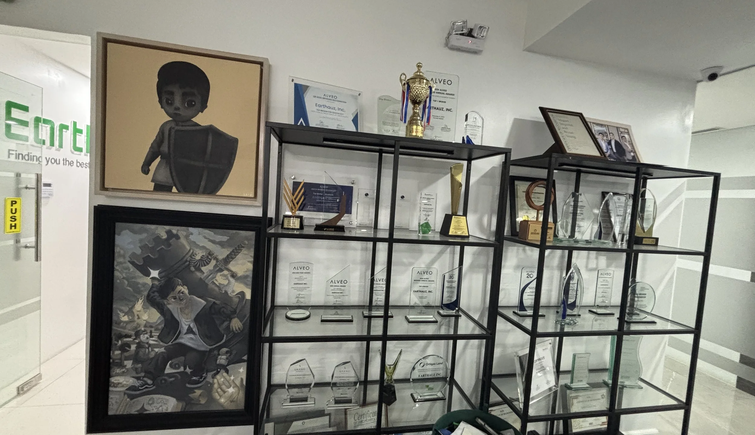 Display of framed artwork, trophies, and multiple glass awards on black metal shelves, with some framed photos and certificates on top.
