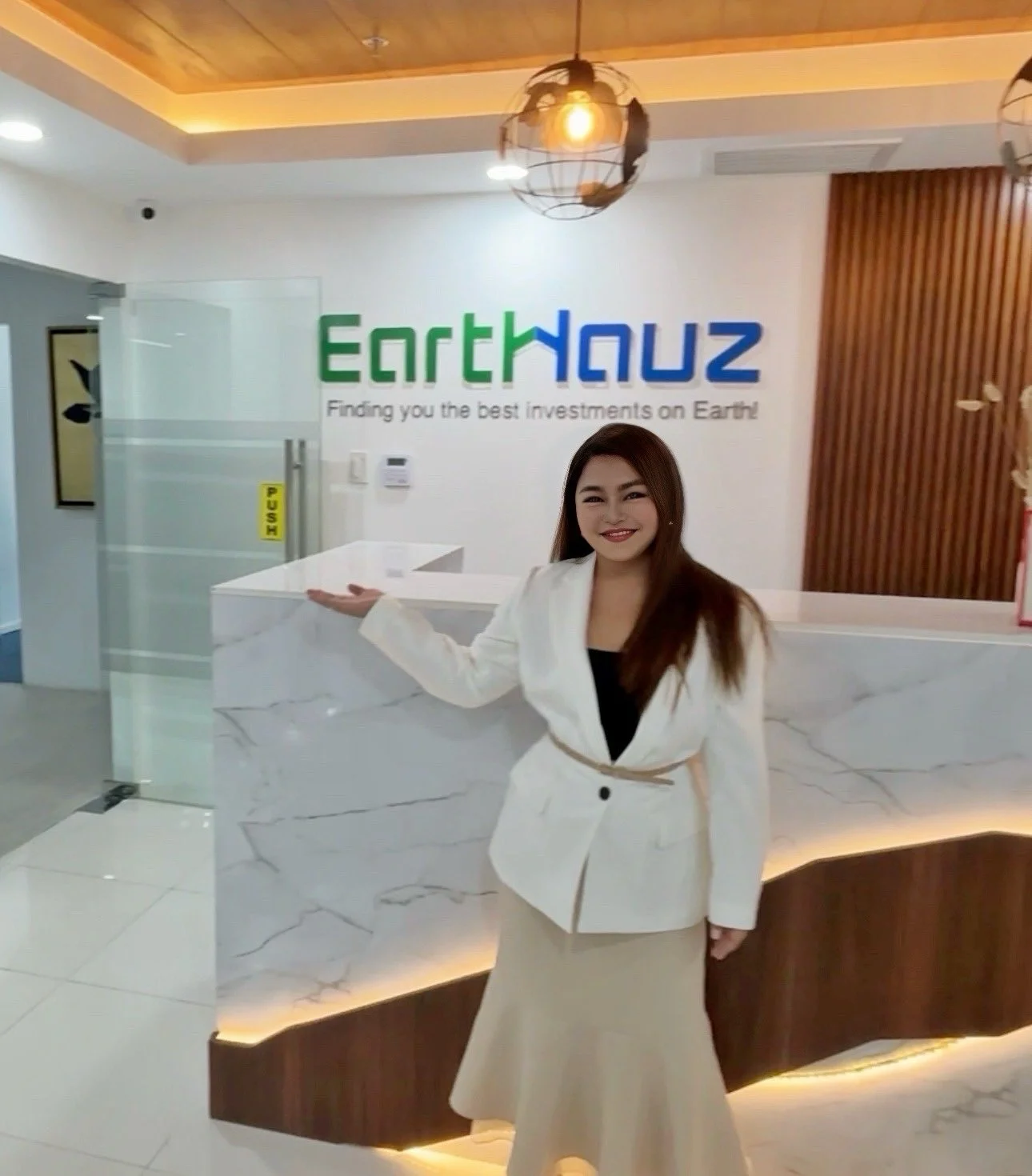 A woman standing at a reception desk in front of a sign that reads 'EarthHauz' with the slogan 'Finding you the best investments on Earth!'. She is smiling, dressed in a white coat and black top, inside an office or corporate environment.