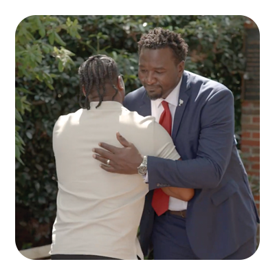 Two men are embracing outdoors with lush green trees and a brick wall in the background.