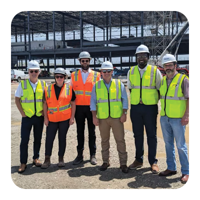 Six people in safety vests and construction helmets standing on a construction site.