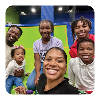 Group of six smiling people, including children and adults, in a colorful indoor play area with a green and blue background.