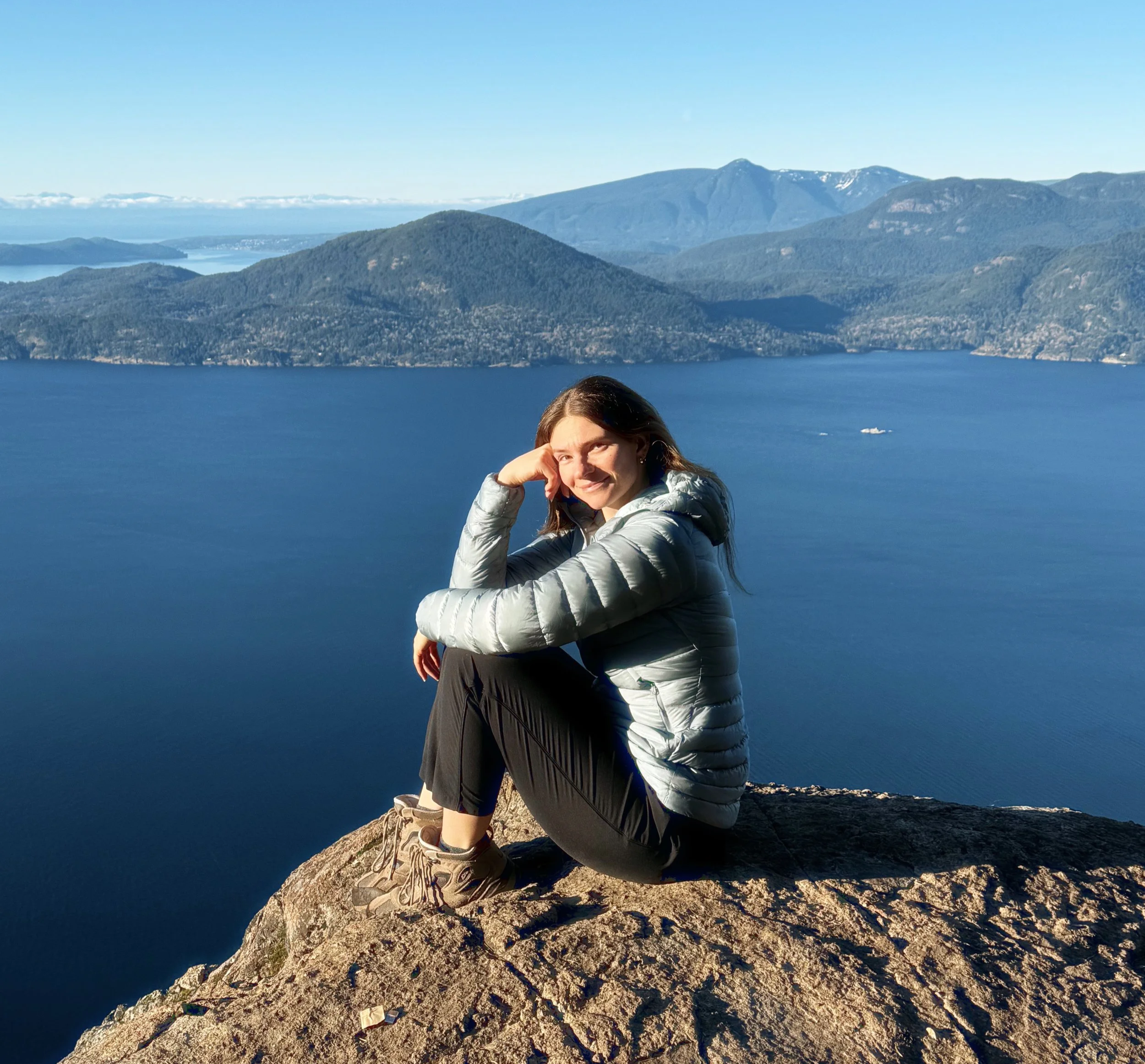 A woman with long brown hair, wearing hiking boots, sitting on a rocky cliff edge with a scenic view of a mountain, a lake, and forested hills in the background.