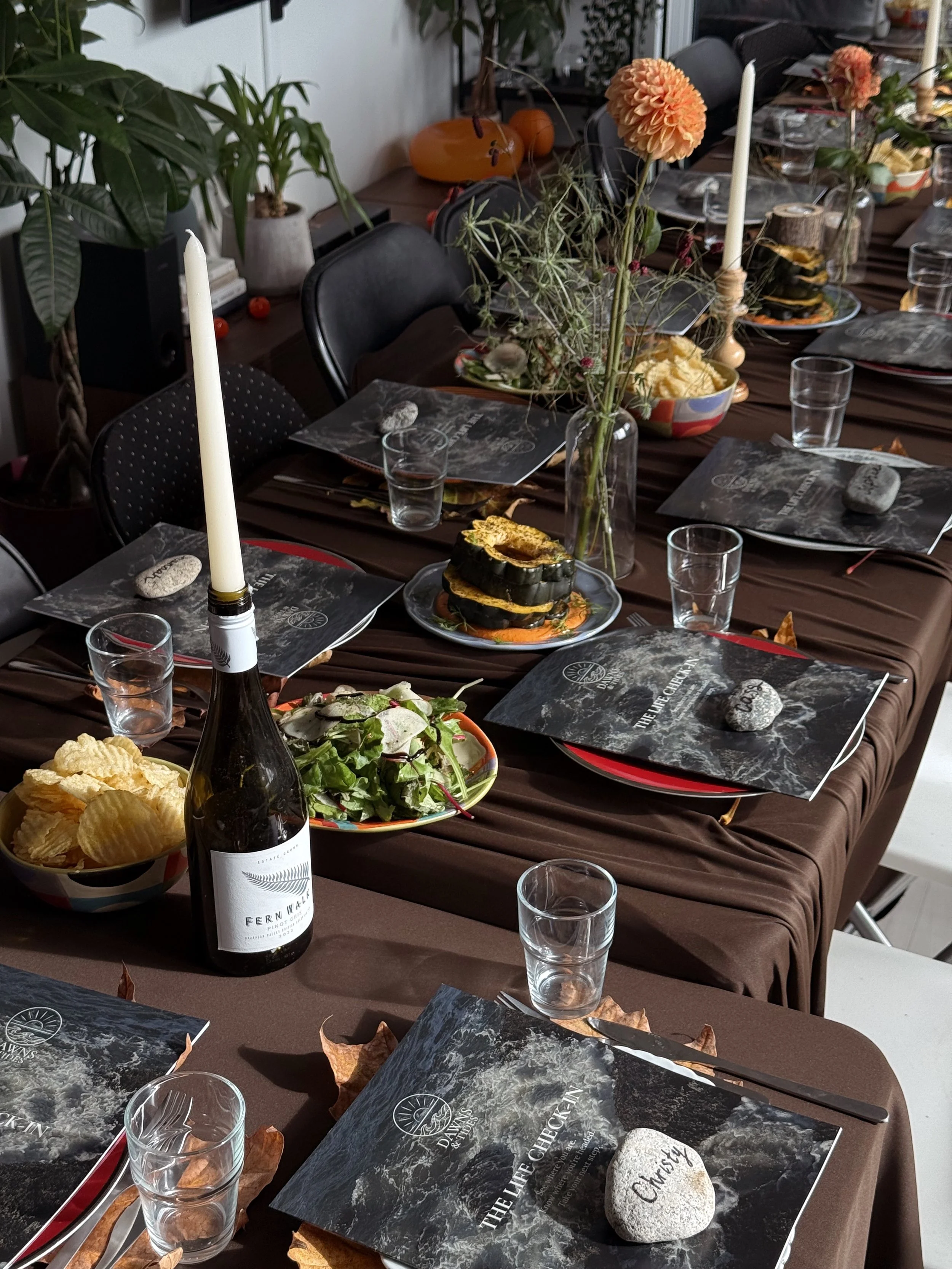 A dining table set for a meal with black and red placemats, plastic water glasses, a bottle of wine, and a Christmas-themed stone placed on each placemat. The table is decorated with tall white candles in candle holders, vases with dried flowers, and bowls of potato chips and salad. In the background, there are potted plants, pumpkins, and other decorations.