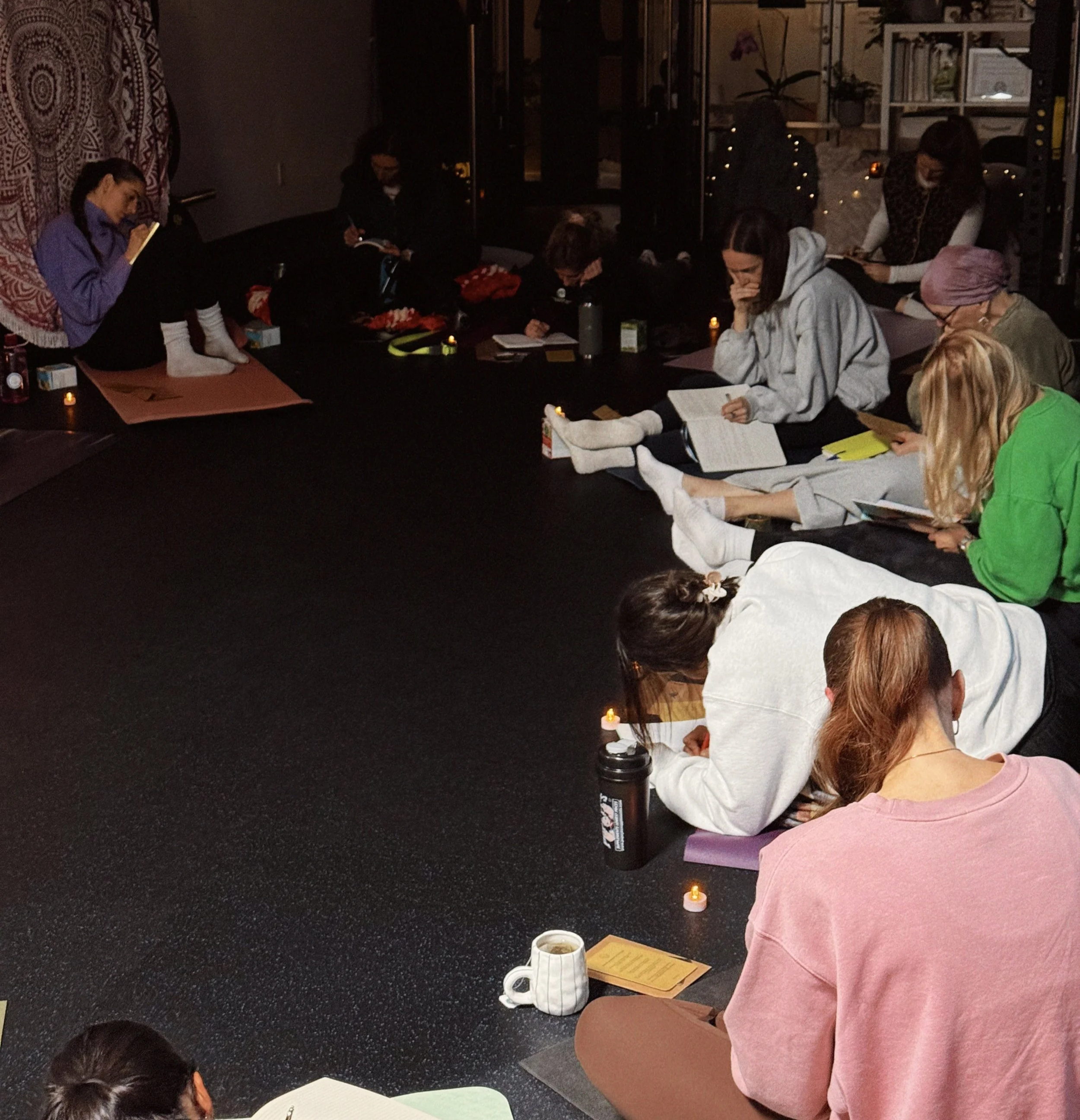 People sitting cross-legged on mats and blankets, praying or meditating in a dimly lit room with candles, books, and decorative wall hangings.