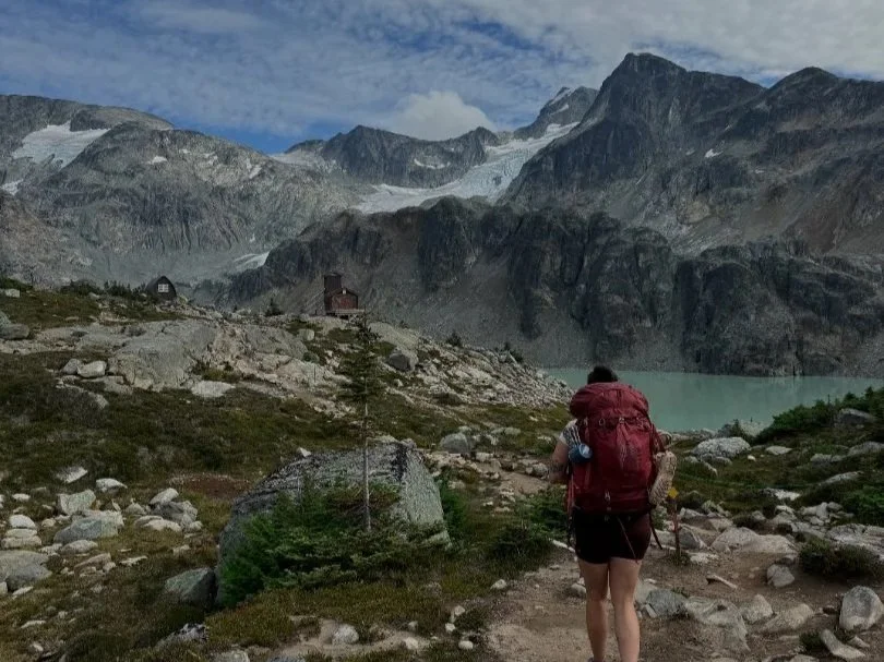 A person hiking through a mountainous terrain with a red backpack, overlooking a lake with rugged scenery and snow-capped peaks in the background.