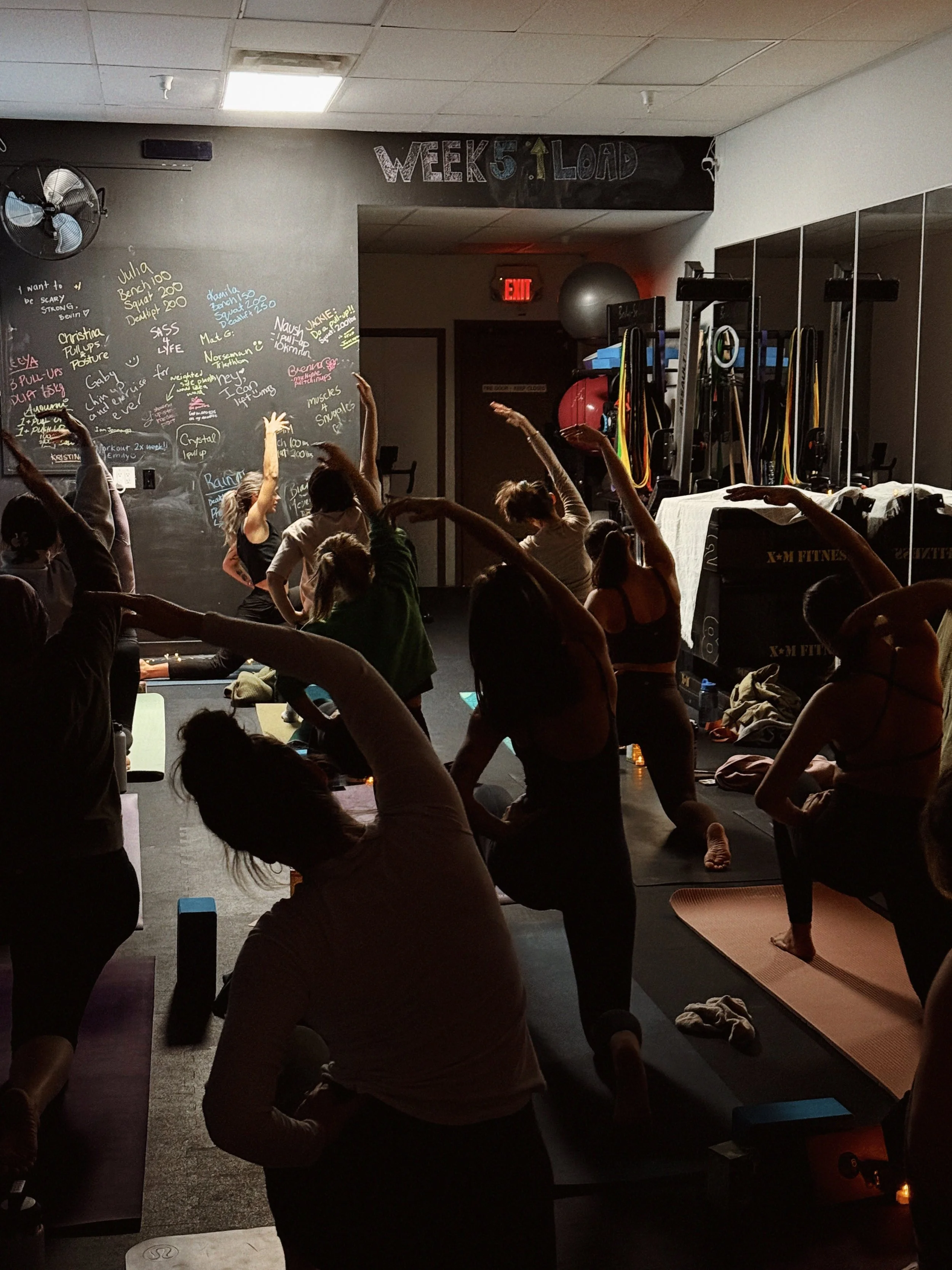 A group of people practicing yoga or stretching exercises in a dimly lit fitness studio with dark walls, some of them are doing stretches with raised arms, while others are in different poses on yoga mats. There is a black chalkboard wall with handwritten notes and names, a fan on the wall, and various fitness equipment including resistance bands and exercise balls.