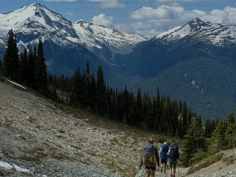 Three hikers walking along a mountain trail with snow-capped peaks, dense forest, and cloudy sky in the distance.