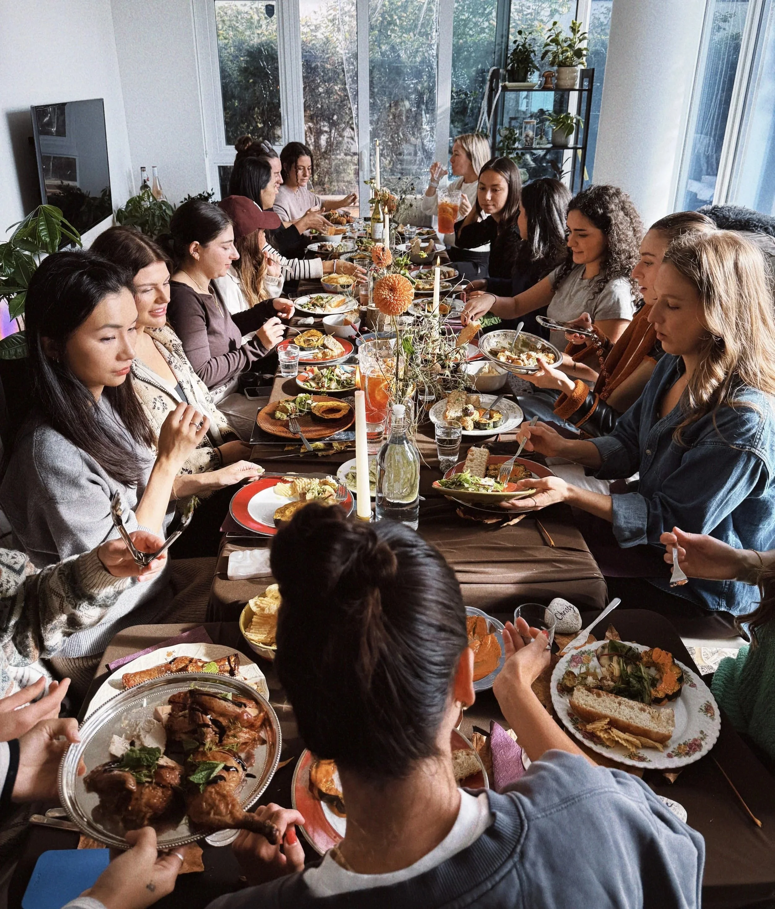 A group of people sitting around a dining table enjoying a meal together in a well-lit room with large windows, plants, and a modern decor.