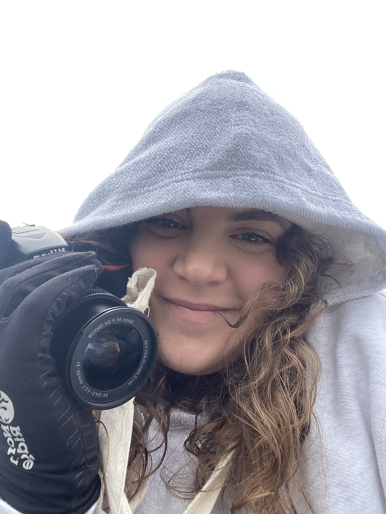 A woman wearing a gray hoodie and black gloves is holding a camera up to her face, smiling with curly hair visible under the hoodie.
