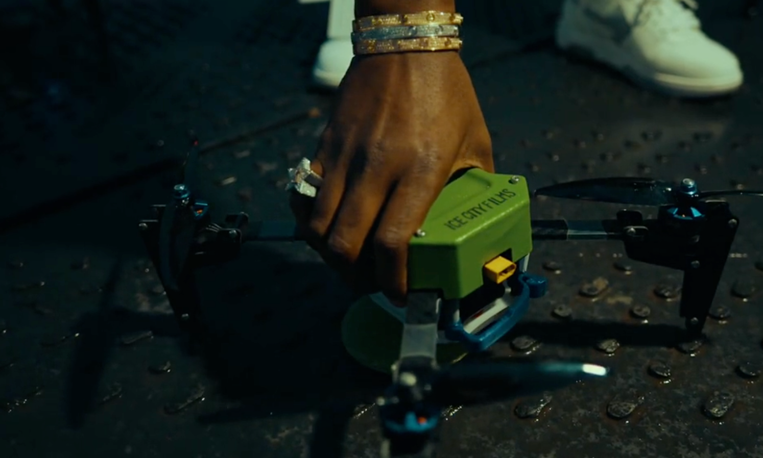 A hand with bracelets and rings reaches down to adjust a green quadcopter drone on a textured dark surface.