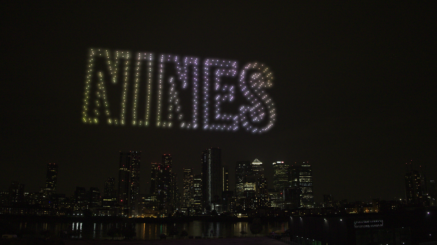 Colorful drone light display spelling 'AMIGOS' in the night sky over a city skyline with illuminated buildings.