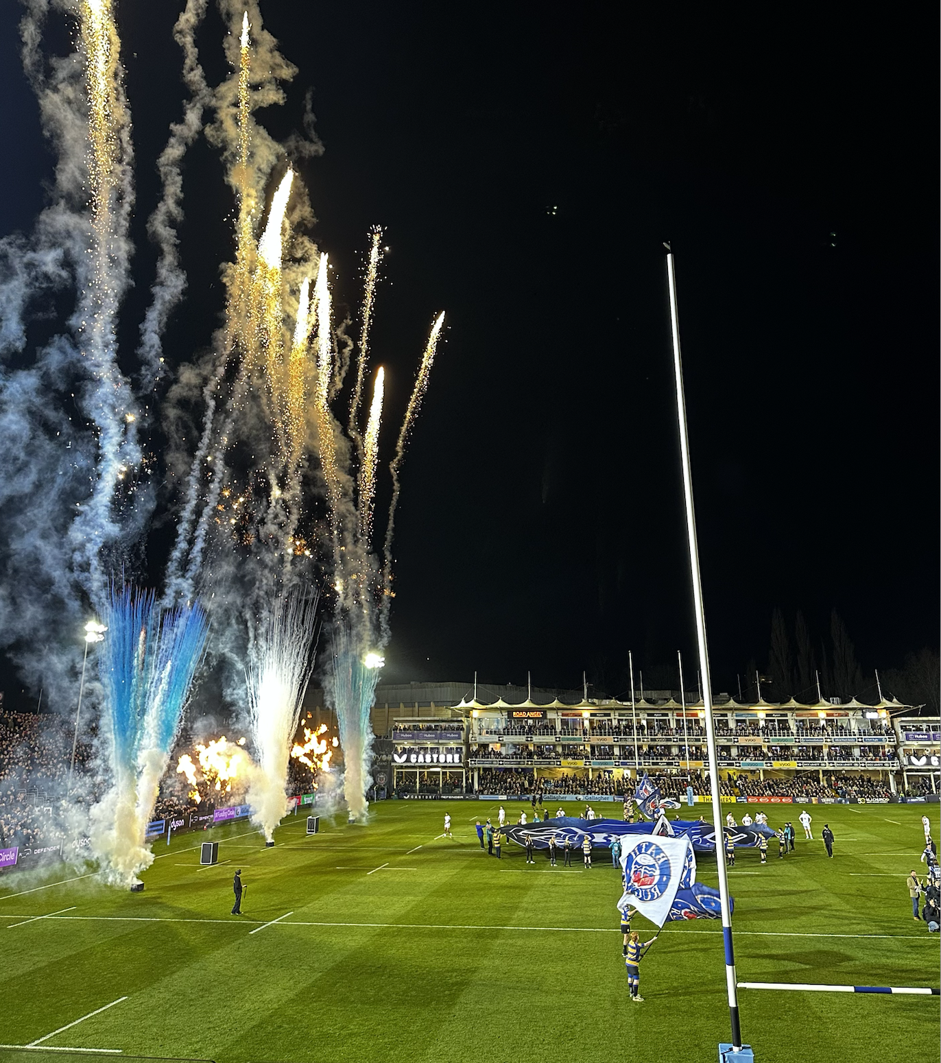 Fireworks display over a rugby stadium during a nighttime match celebration with players and flags on the field.